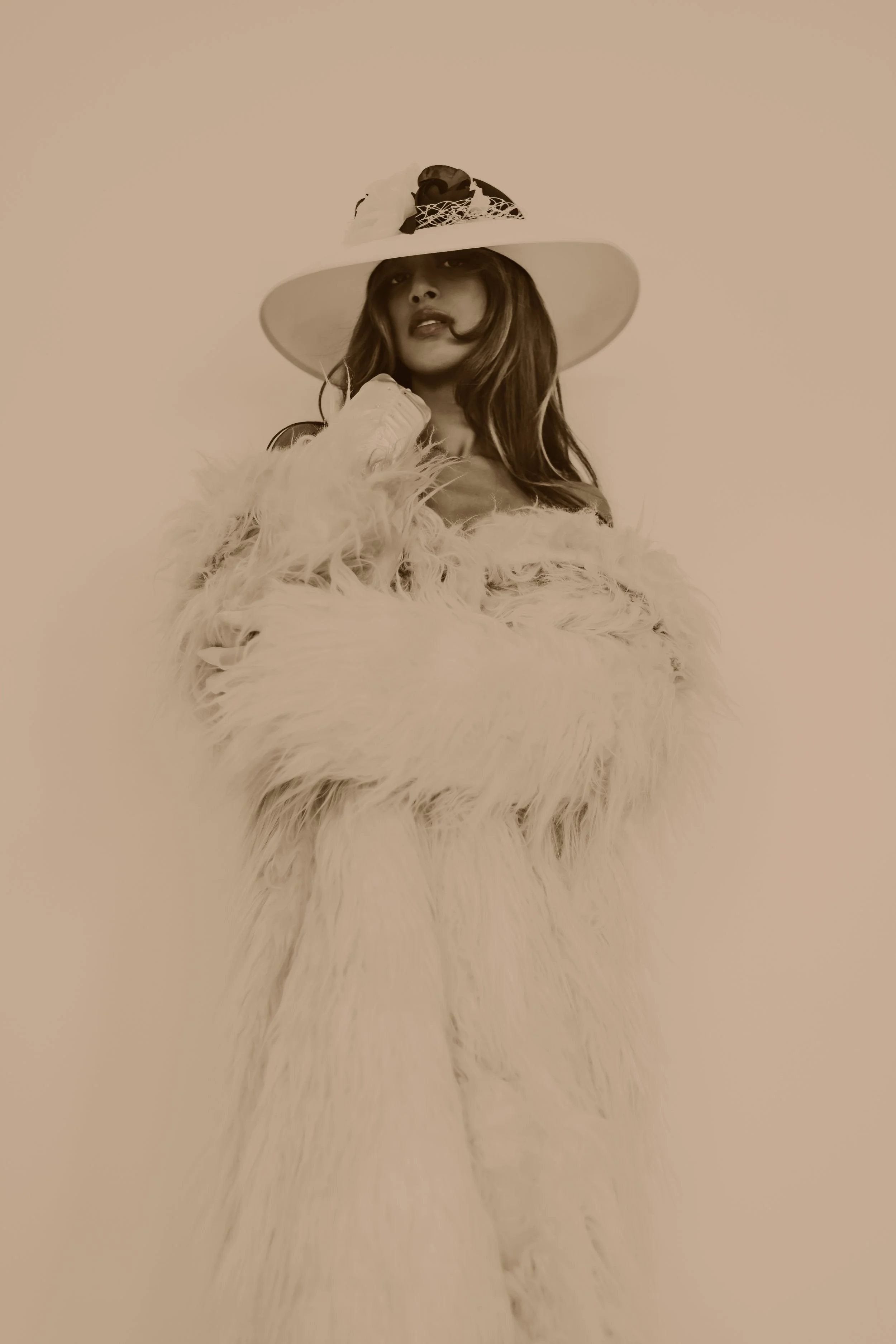 A woman wearing a large hat decorated with flowers and netting, and a fluffy fur coat, posing against a plain background.