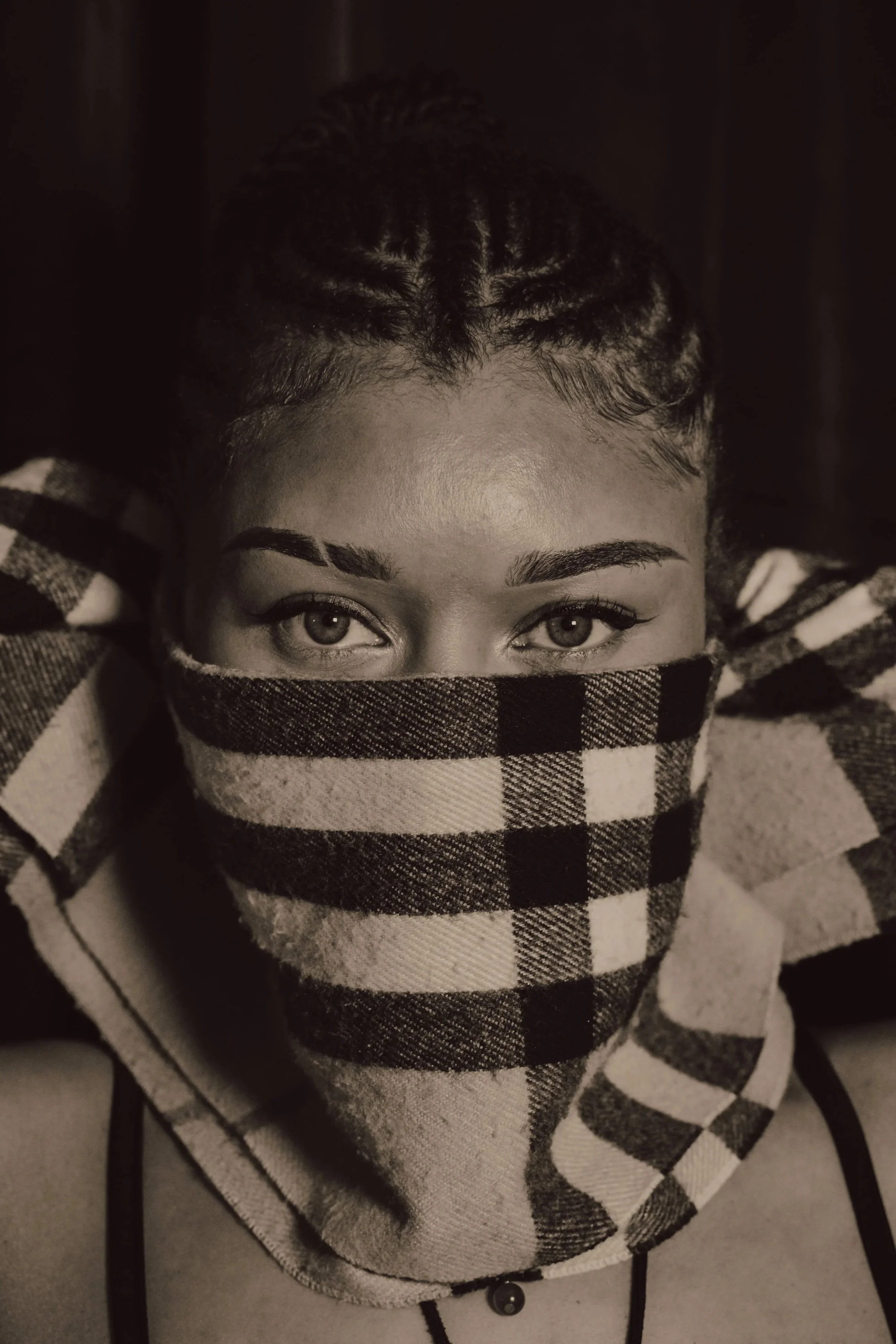 A woman with curly hair and light-colored eyes looking over her shirt, which has a checkered pattern, while wearing a hoodie and a face mask.