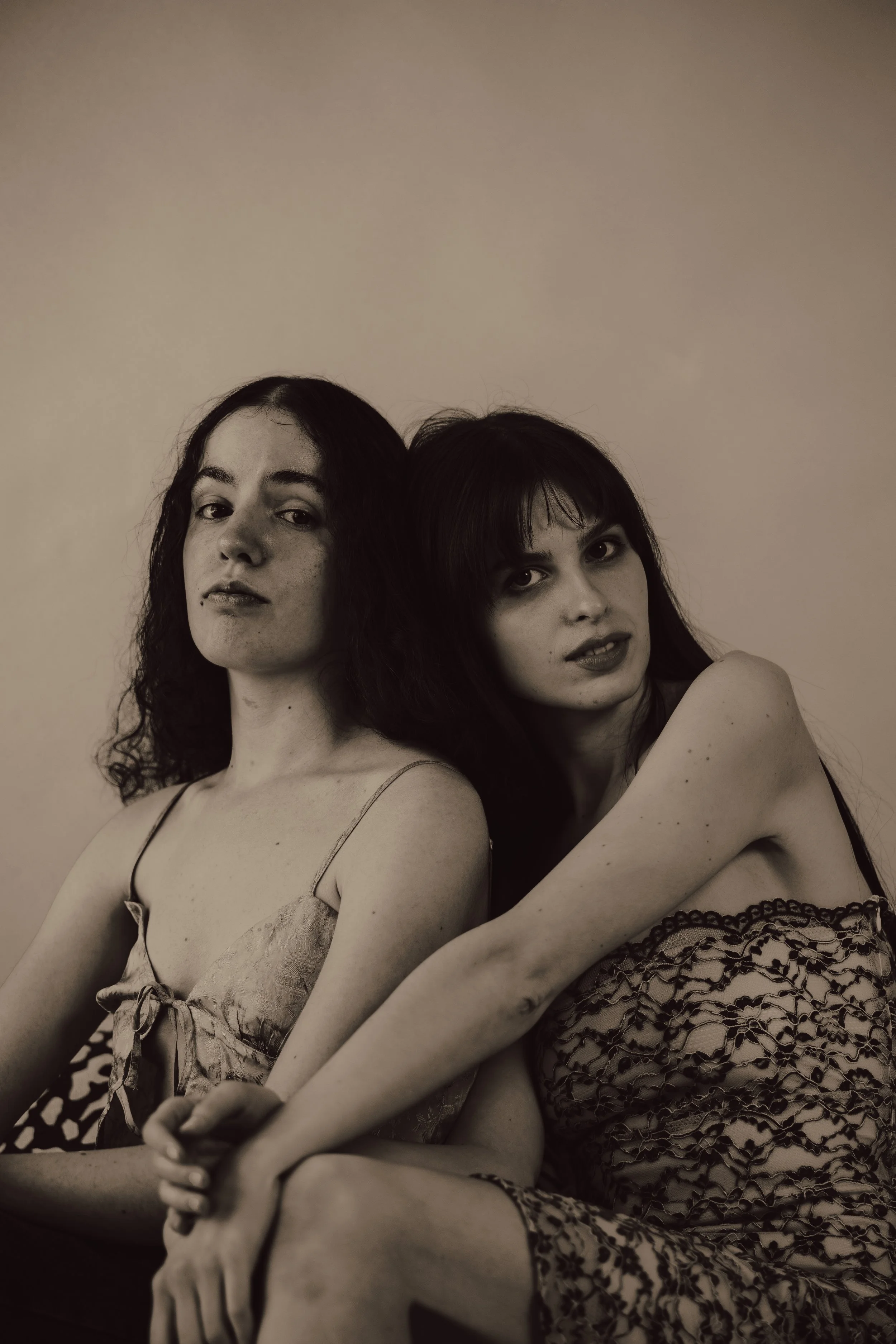 Two women with dark hair and expressive eyes sit close together against a plain light-colored background, wearing patterned dresses.