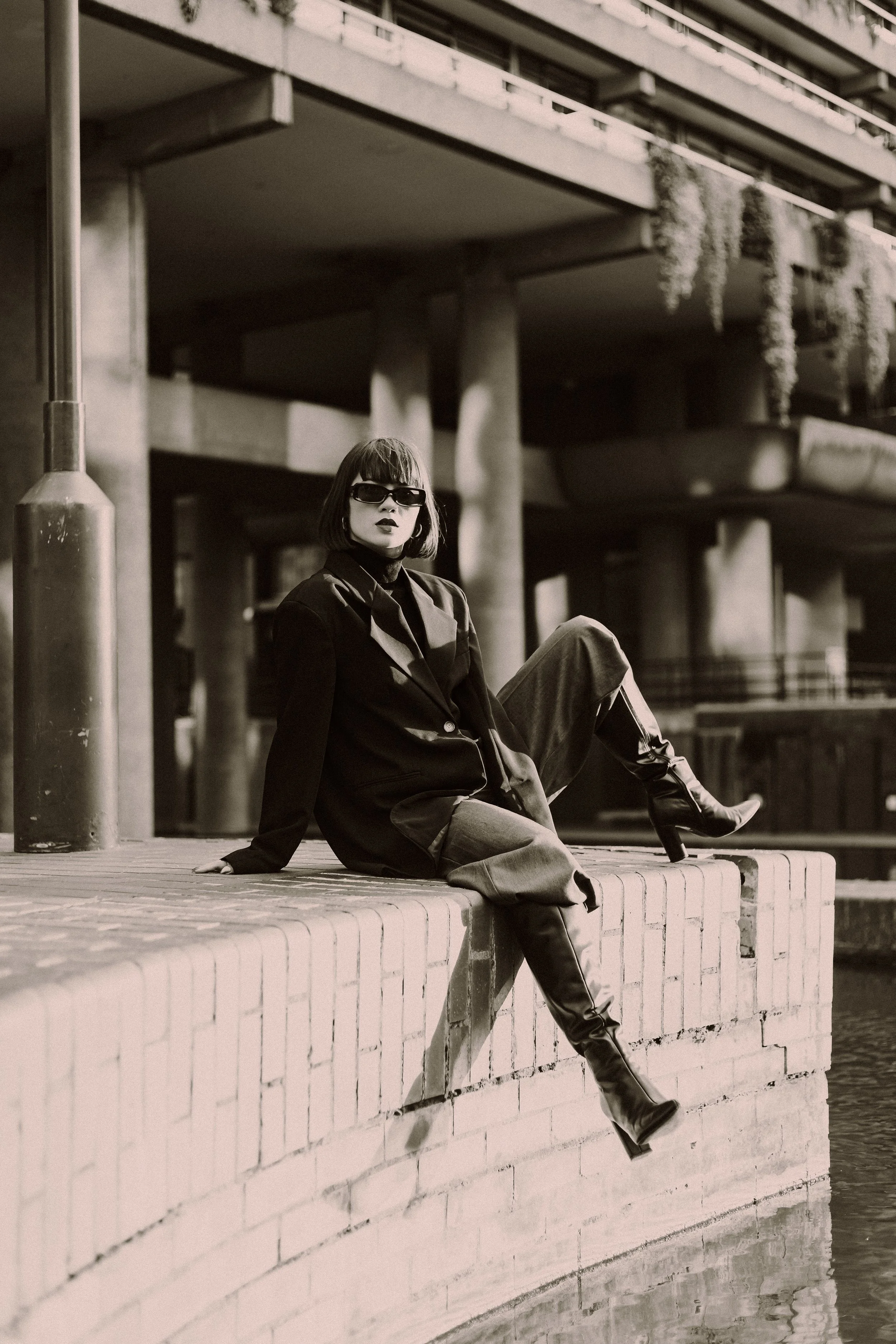 A woman with short hair, wearing sunglasses and a blazer, sitting on a brick ledge under a bridge or overpass, with hanging plants and concrete pillars visible in the background.