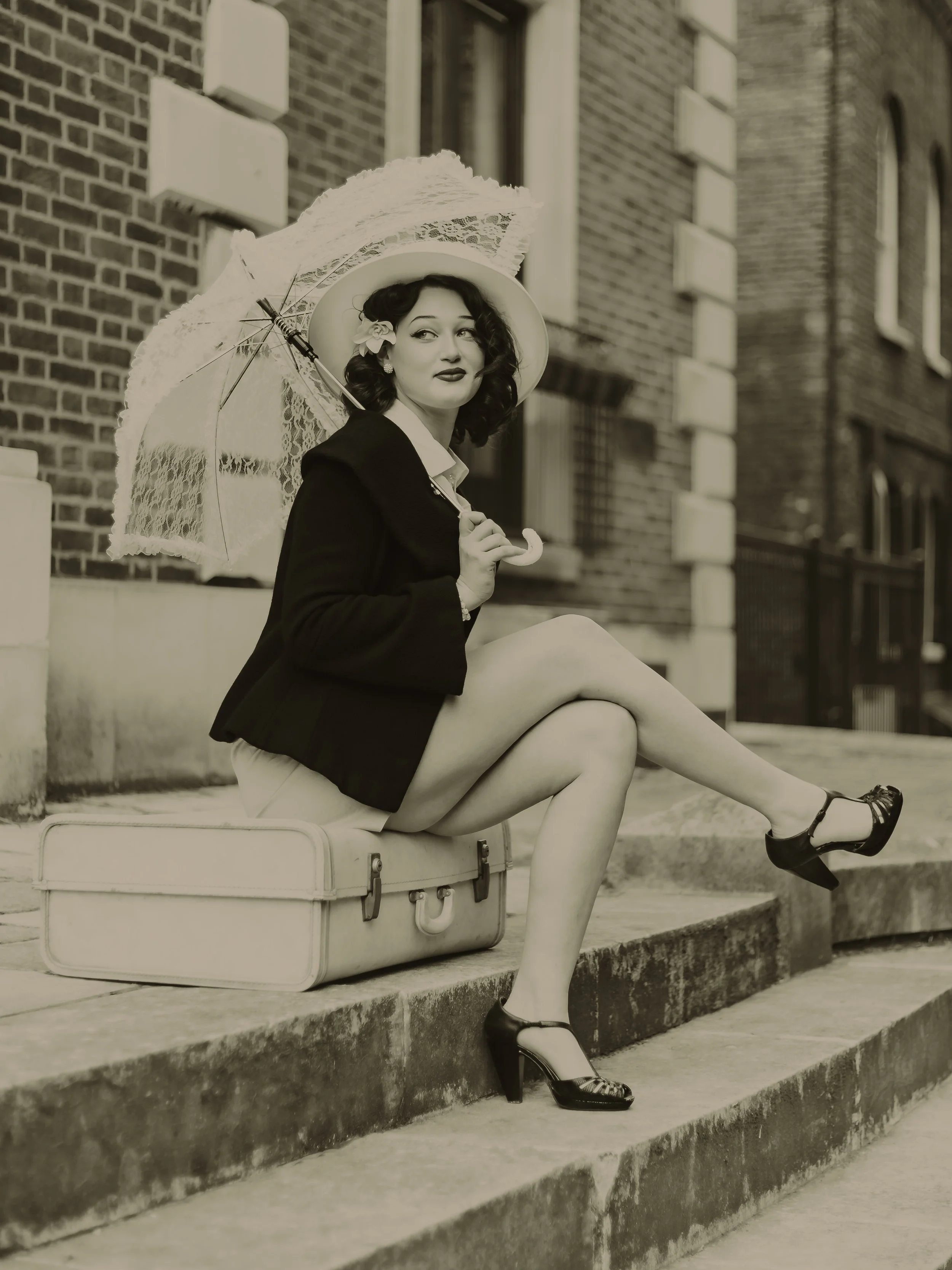 A woman dressed in vintage clothing sitting on a suitcase on the curb, holding a lace parasol, with brick buildings in the background.