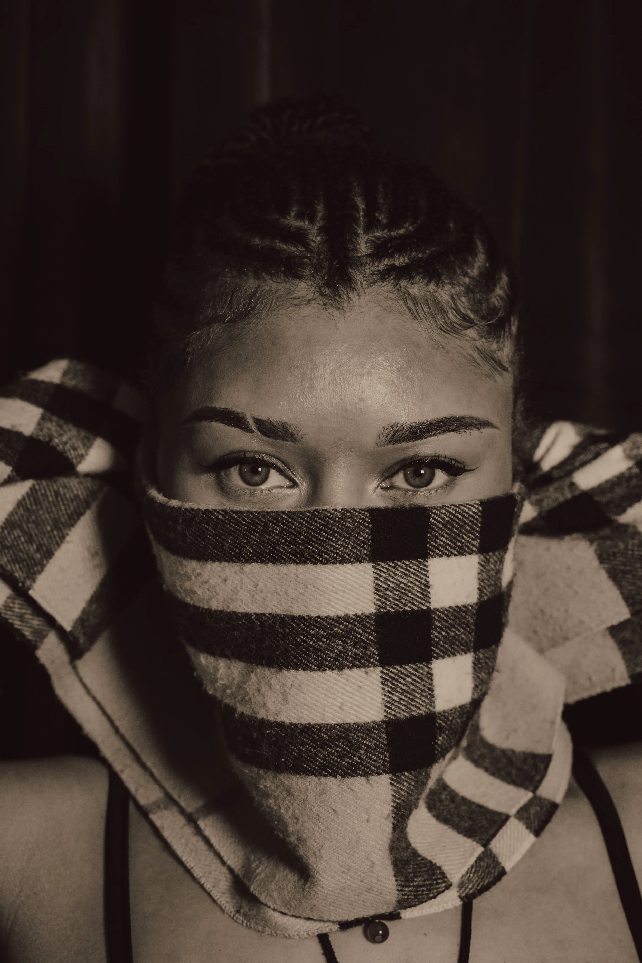 A person wearing a checkered scarf covering their lower face, with only their eyes visible, lying on a cushioned surface.