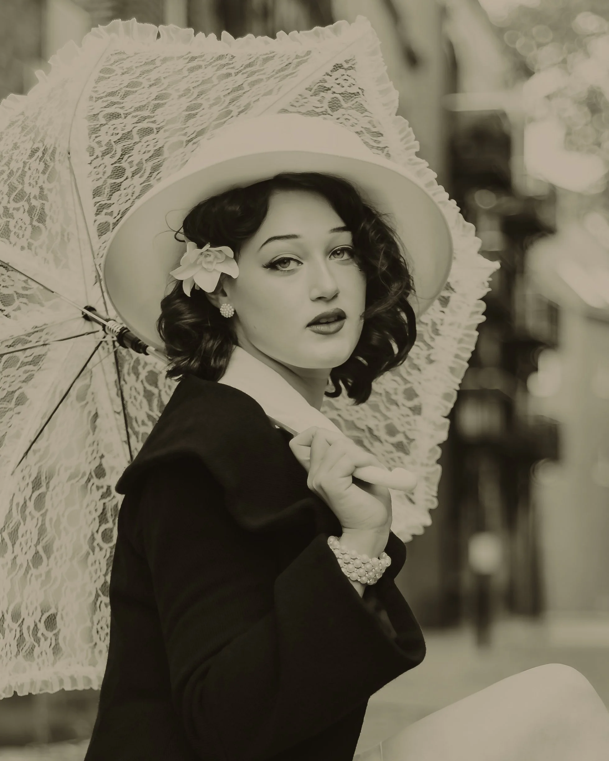 A black and white vintage style photograph of a woman holding a lace parasol. She is wearing a wide-brimmed hat with a flower, earrings, a blazer, and a pearl bracelet, with a confident expression and curly hair.