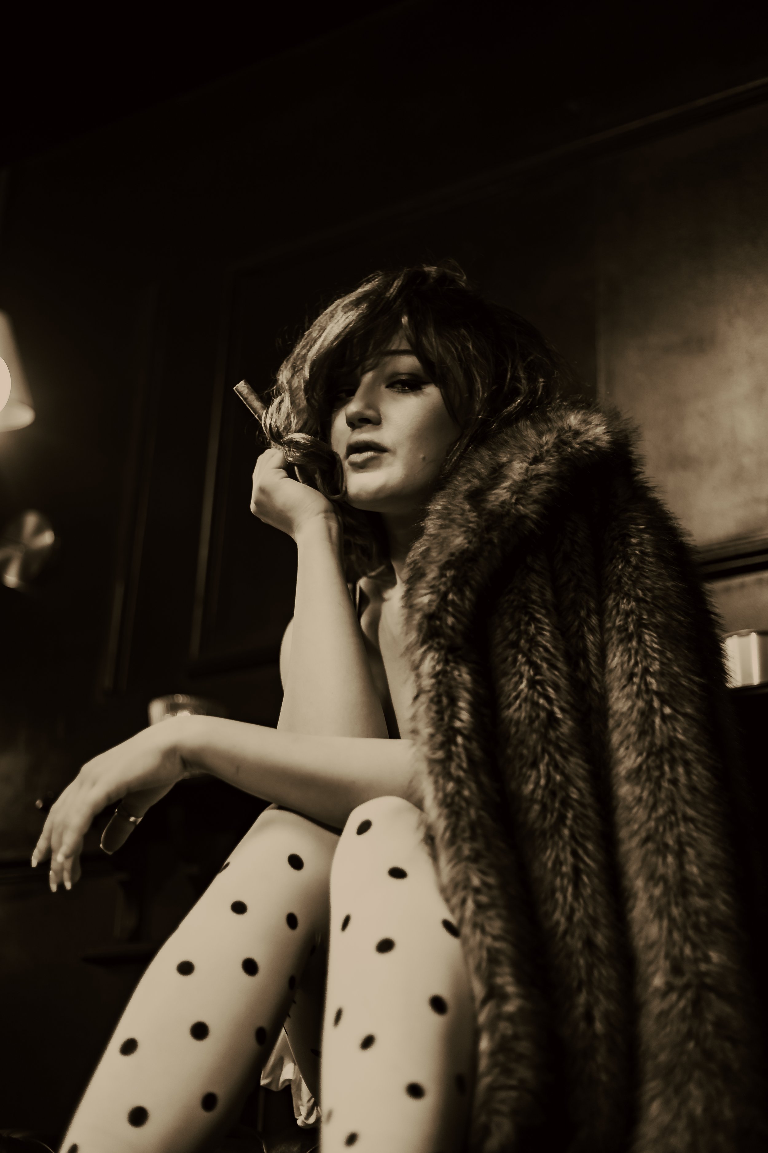 Woman in a fur coat and polka-dot tights smoking a cigar by a window, photographed in warm monochrome