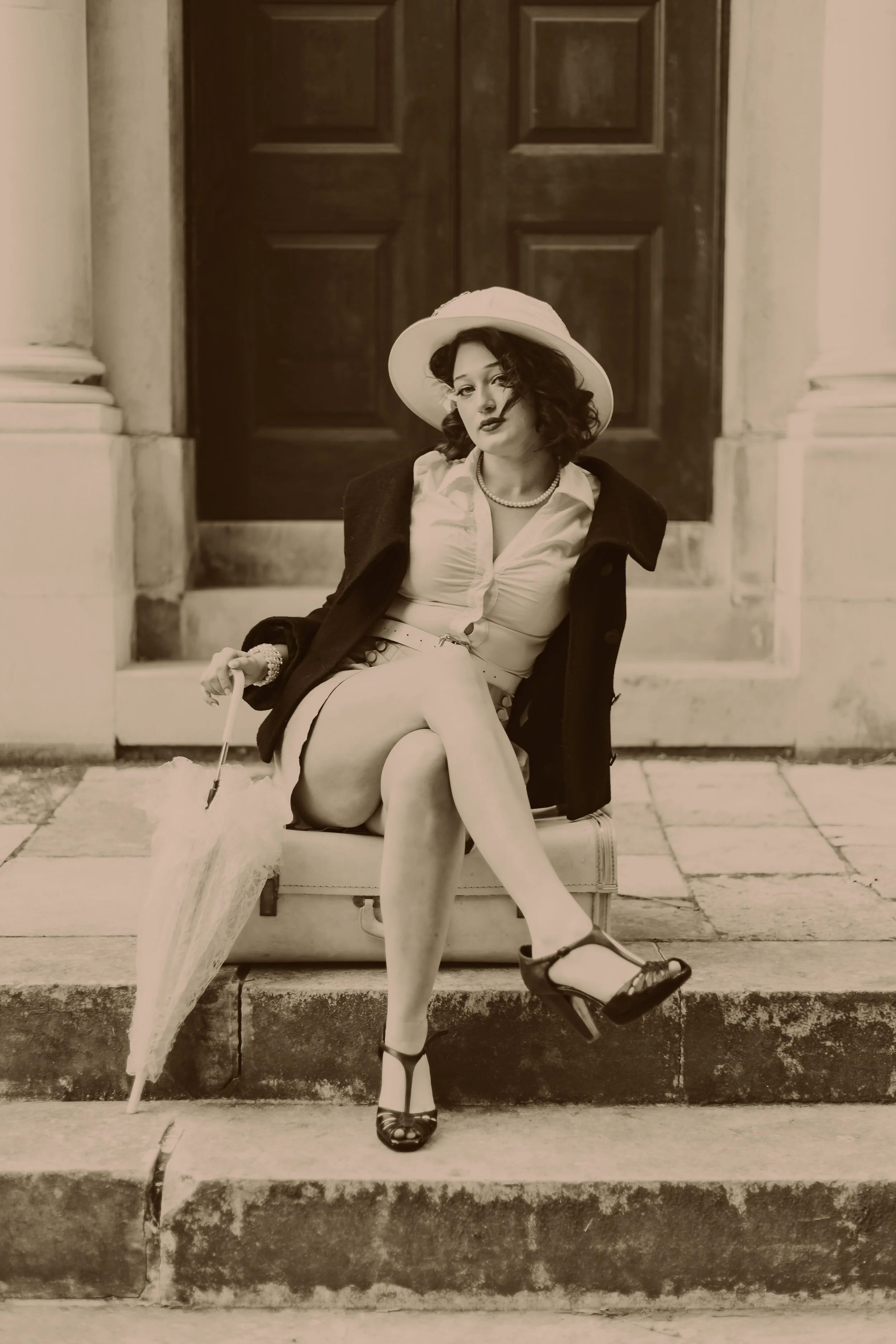 A woman in vintage clothing sitting on a suitcase on steps, holding an umbrella and wearing a hat, coat, blouse, skirt, and heels.