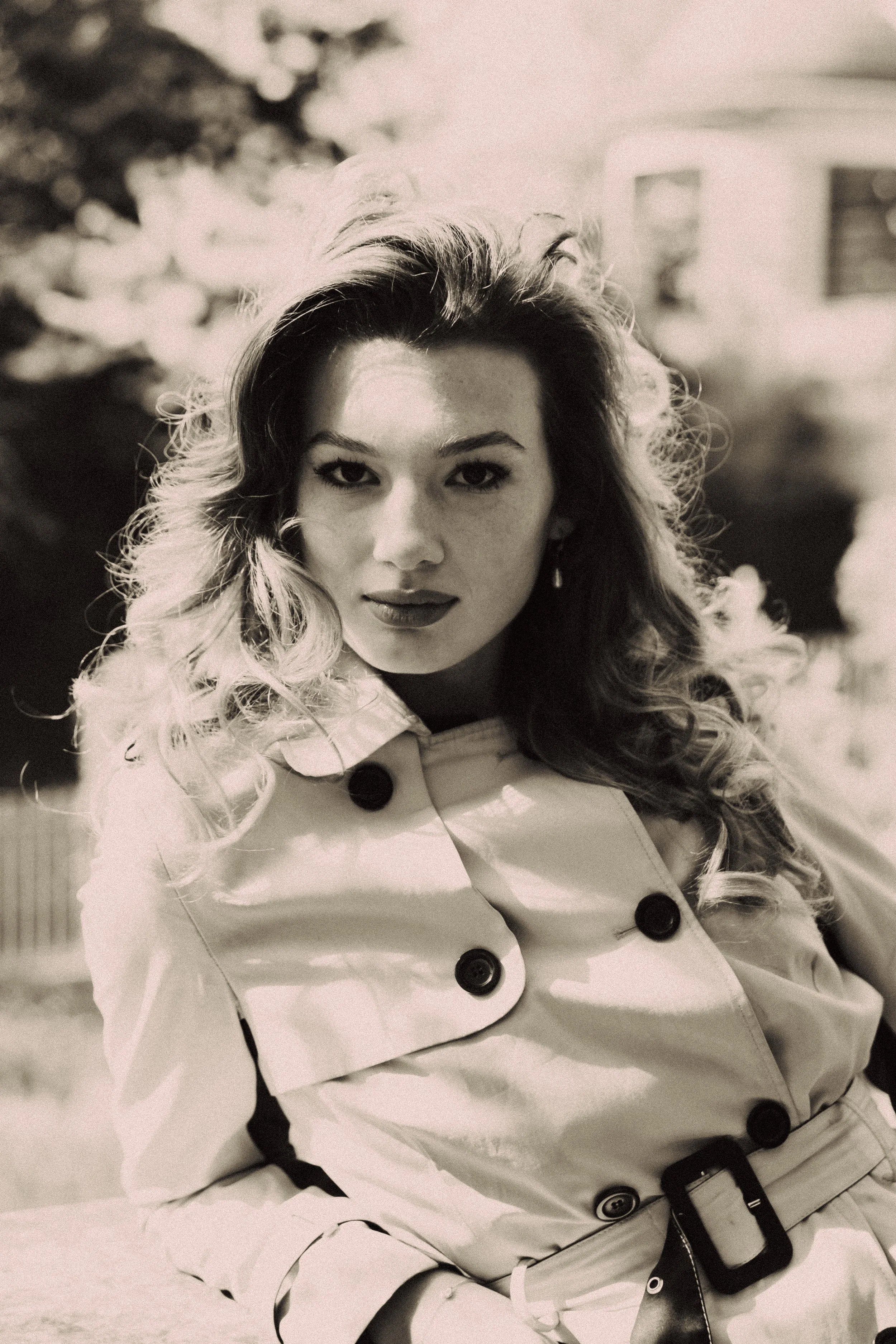 A woman with curly hair wearing a light-colored coat with large buttons, standing outdoors in front of blurred trees and buildings.