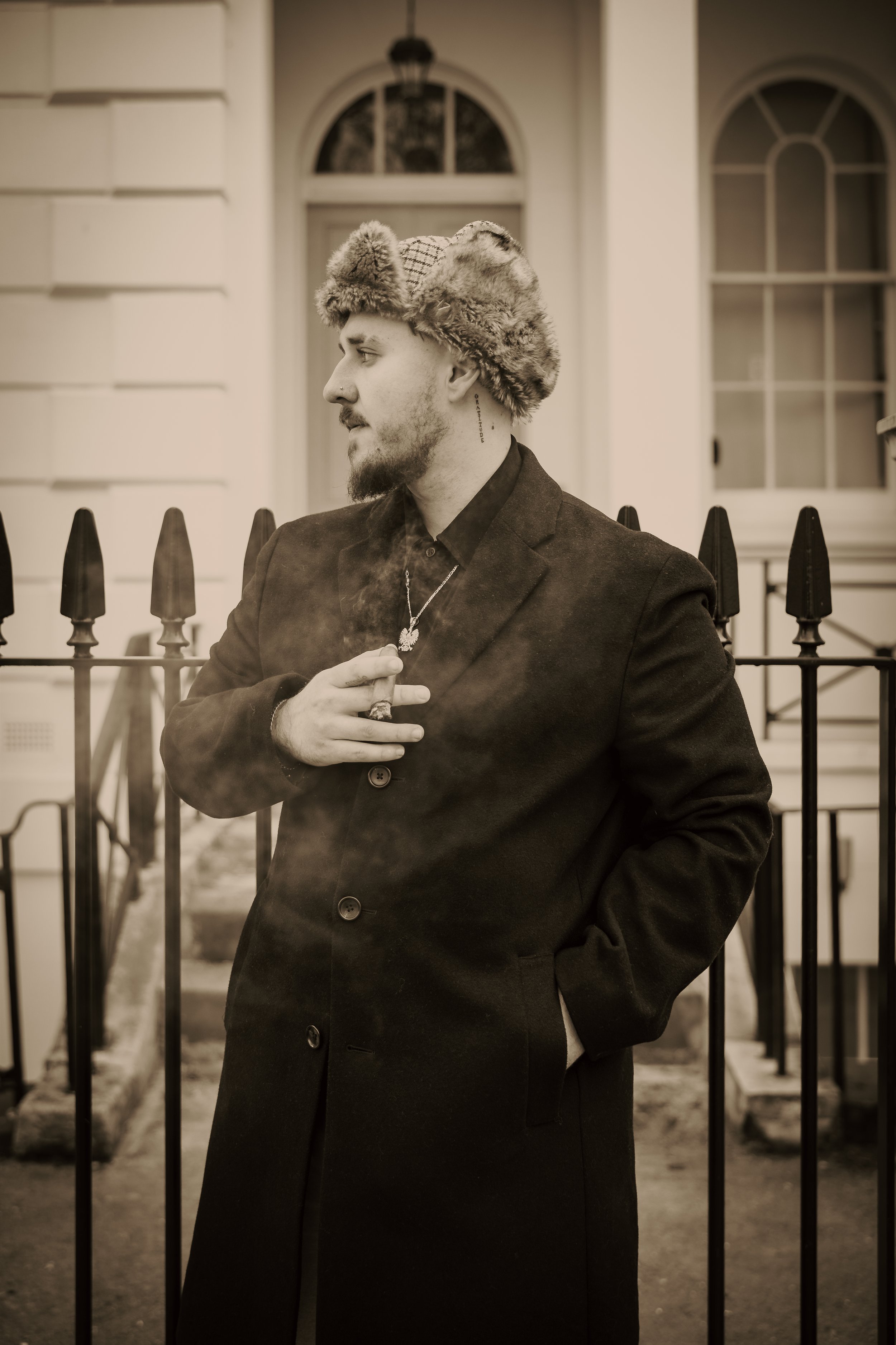 Filip Szczepaniak in a long coat and fur hat outside a Georgian townhouse in London, in sepia monochrome