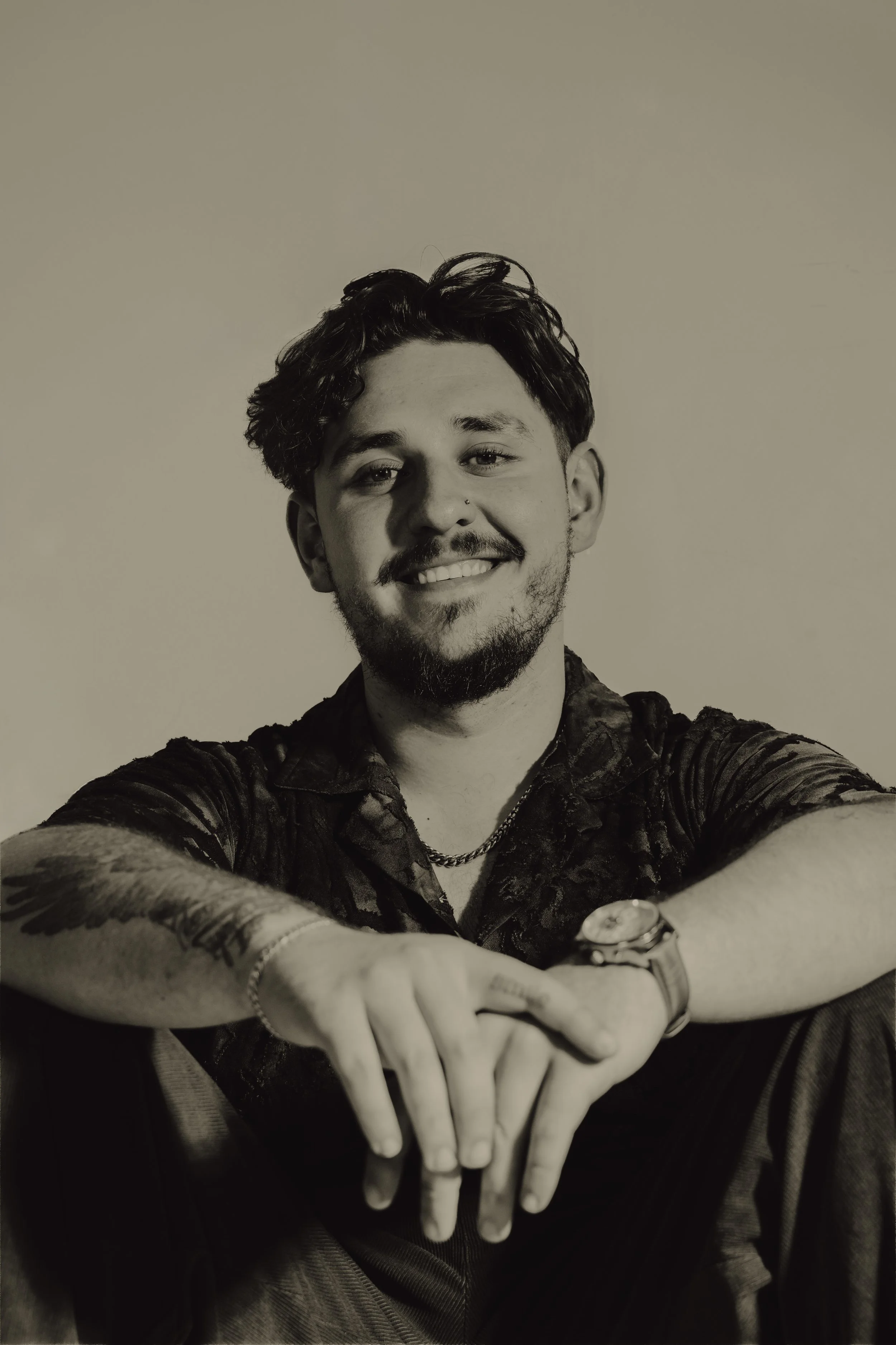 A young man with dark wavy hair and a beard smiling and pointing towards the camera. He is wearing a patterned shirt, a watch, and accessories. The background is plain, and the photo is in black and white.
