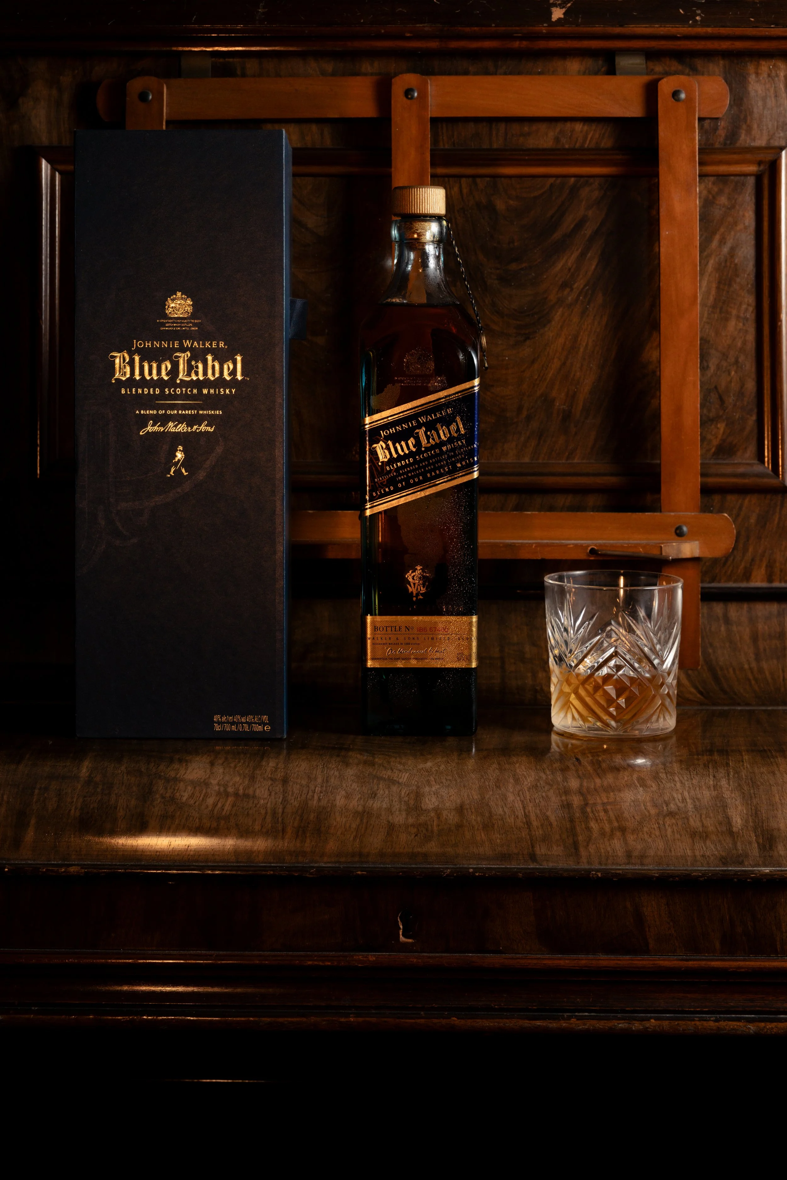 Whisky bottle and its presentation case on a polished wooden bar rack beside a cut-crystal tumbler, lit by a single warm lamp