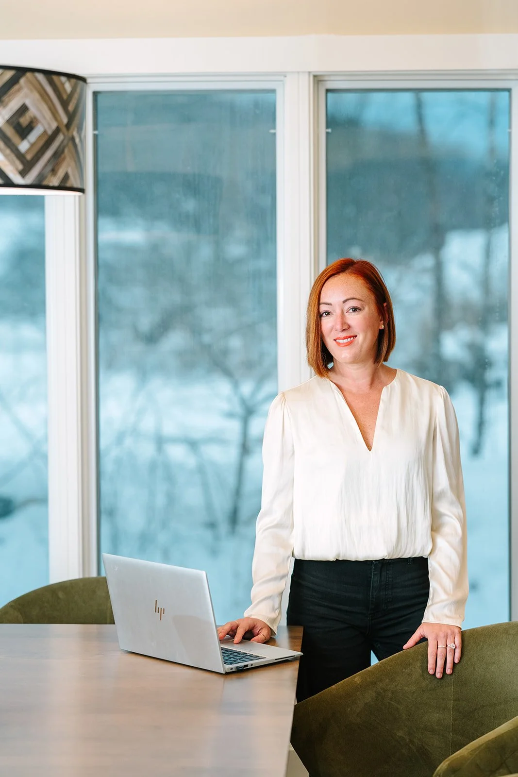 A woman with red hair standing in a modern office space next to a laptop on a wooden table, with large windows showing an outdoor scene with trees in the background.