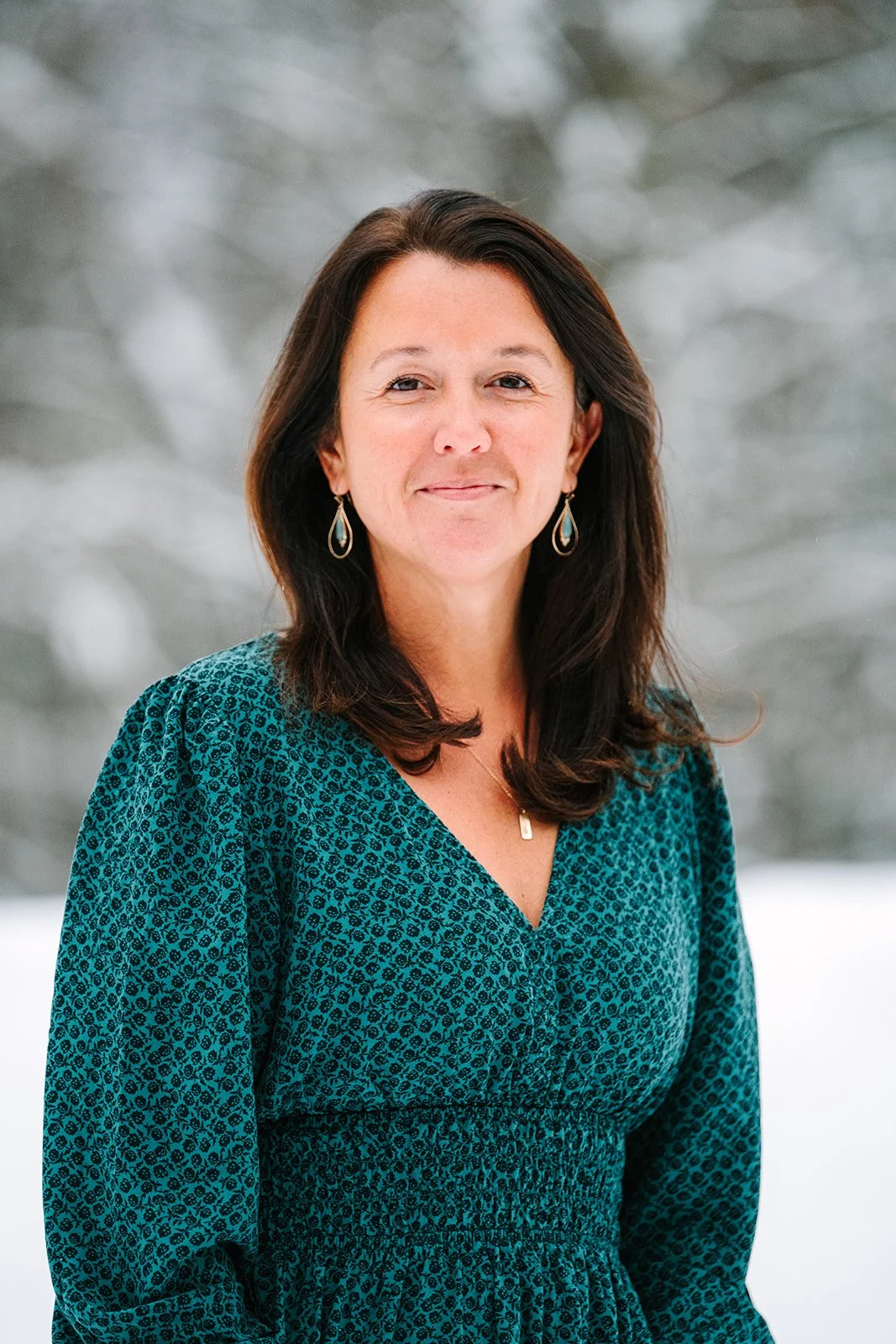 A smiling woman with dark brown hair, wearing a teal patterned dress, earrings, and a necklace, stands outdoors in a snowy landscape with blurred trees in the background.