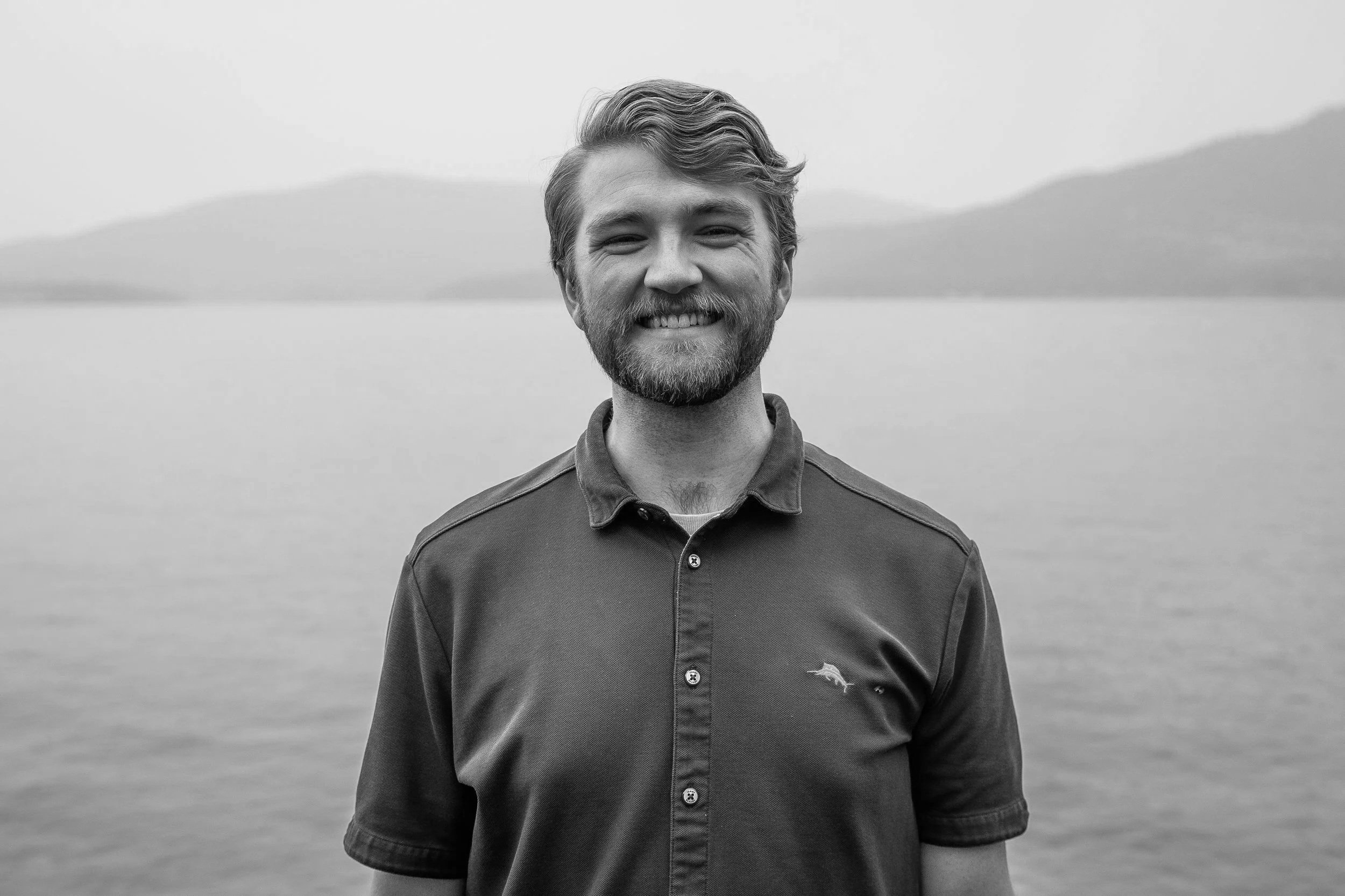 Black and white photo of a smiling young man with short hair, wearing a plaid button-up shirt, standing in front of dark curtains.