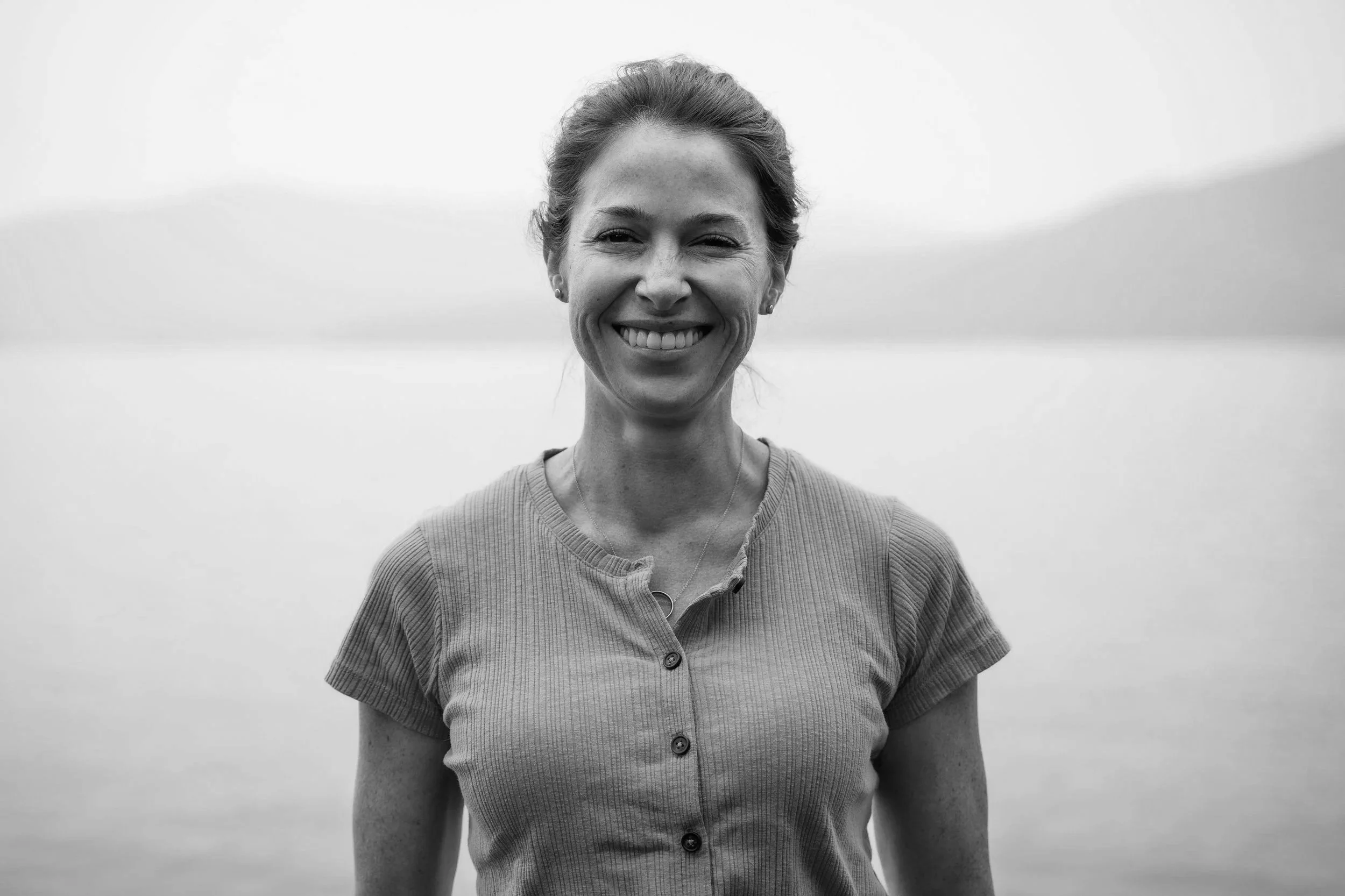 Black and white photo of a smiling woman with long hair wearing a striped shirt, standing in front of a dark curtain.