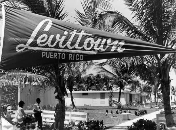Beach scene in Puerto Rico with palm trees, a large Levitown umbrella, and several people near a white building.