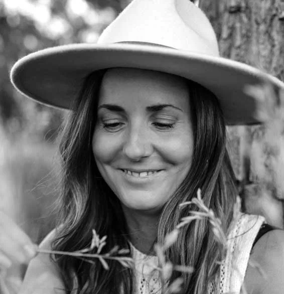 A woman with long hair and a hat smiling as she looks at a sprig of plant she is holding, outdoors near a tree.