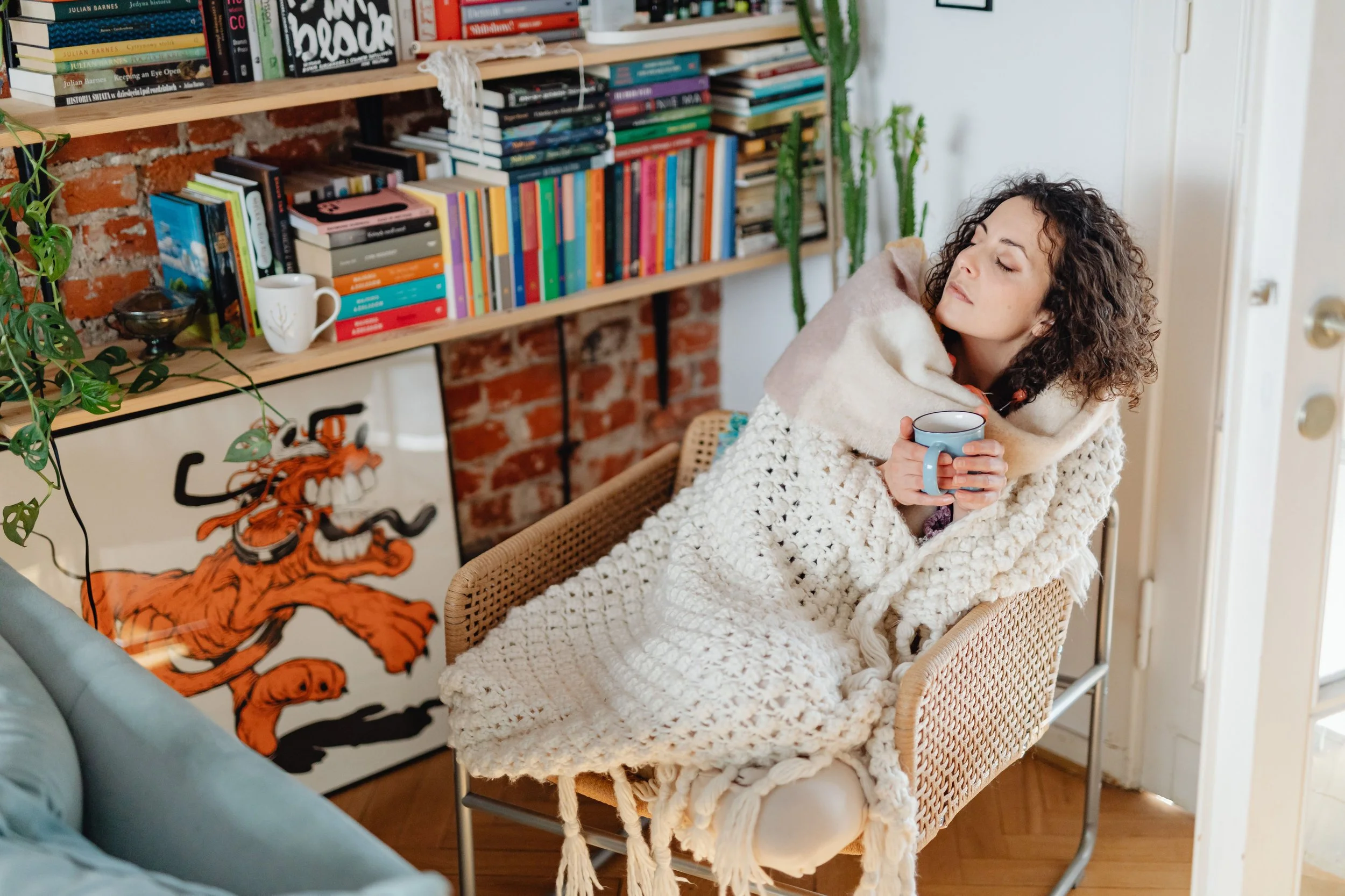 eyes closed a woman sitting peacefully on a chair covered by cozy blankets and holding a mug