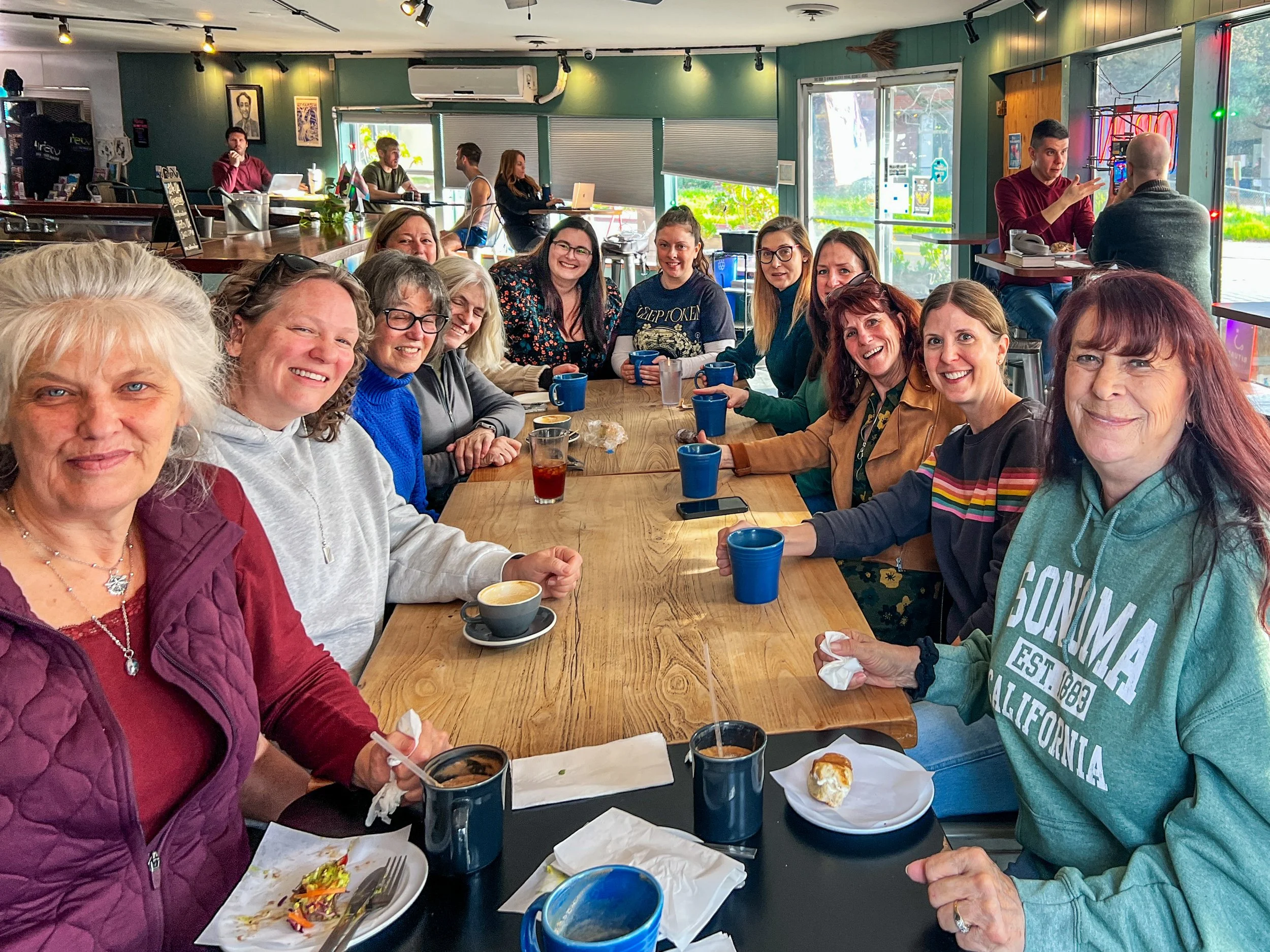 A group of women sitting around a long wooden table in a coffee shop, smiling at the camera, with coffee cups and plates of food on the table.
