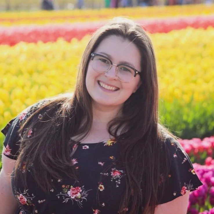 Alex Hailey, a woman with long brown hair and glasses smiling outdoors in a field of colorful tulips.