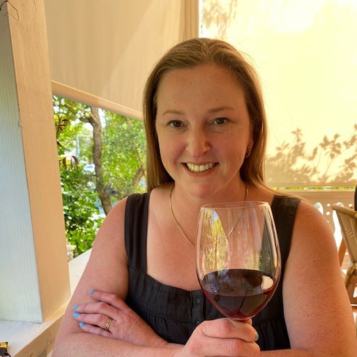 Amanda Garland, a woman smiling and holding a glass of red wine indoors near a window with green trees outside.