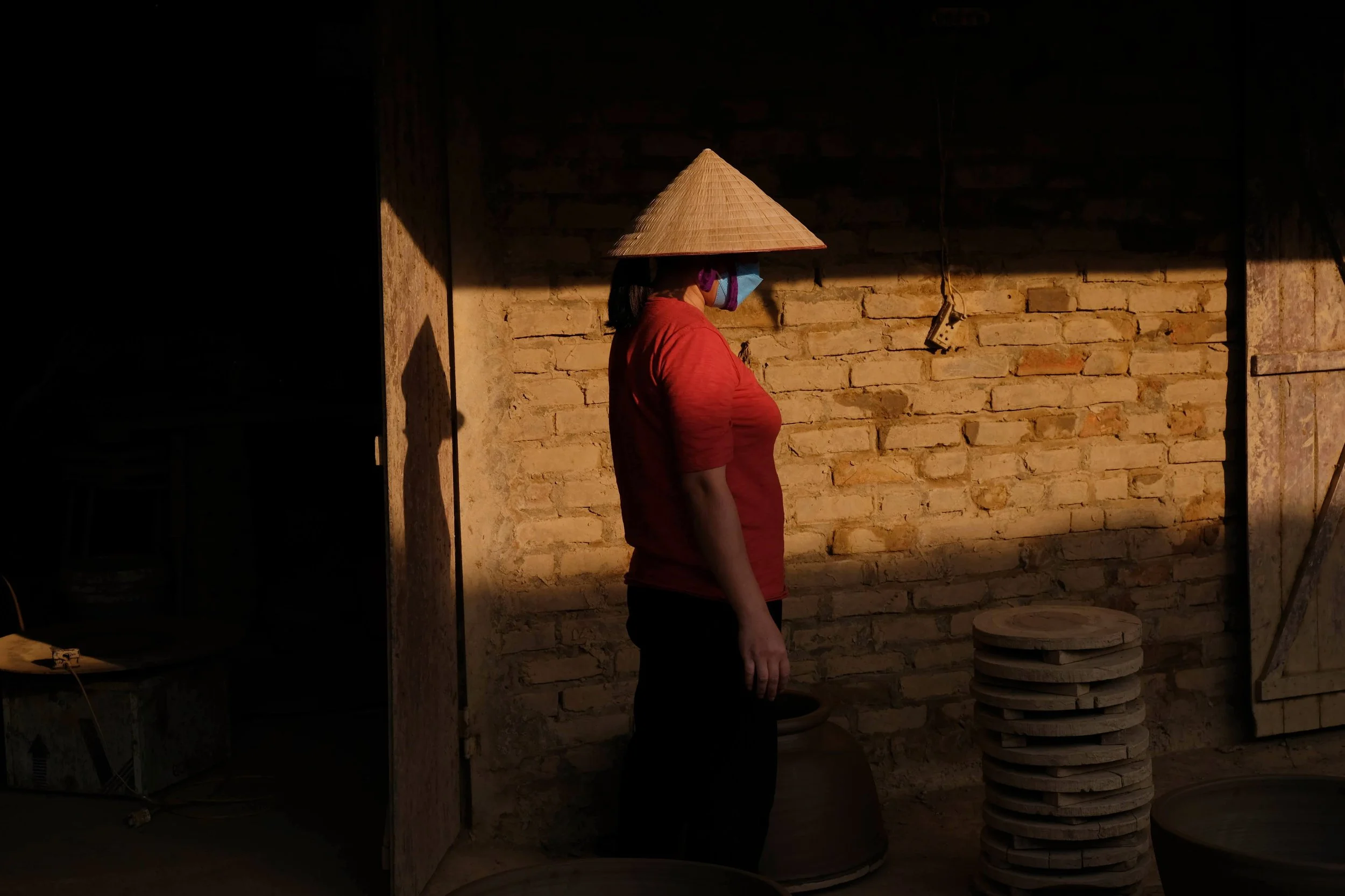 A woman wearing a conical hat and a red shirt stands indoors against a brick wall, with sunlight casting a shadow on her.