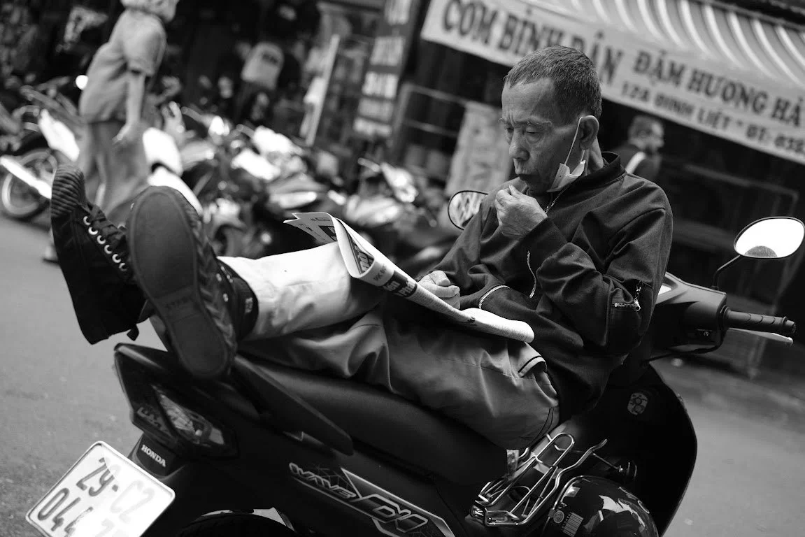 A man sitting and lying on a motorcycle on the street, reading a newspaper, with a face mask pulled down below his chin.