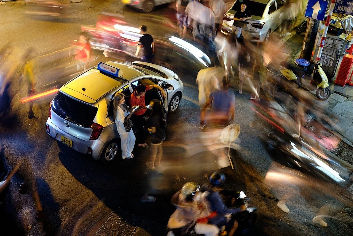 Nighttime city street scene with a yellow taxi and a white car surrounded by blurred moving pedestrians, cyclists, and vehicles.