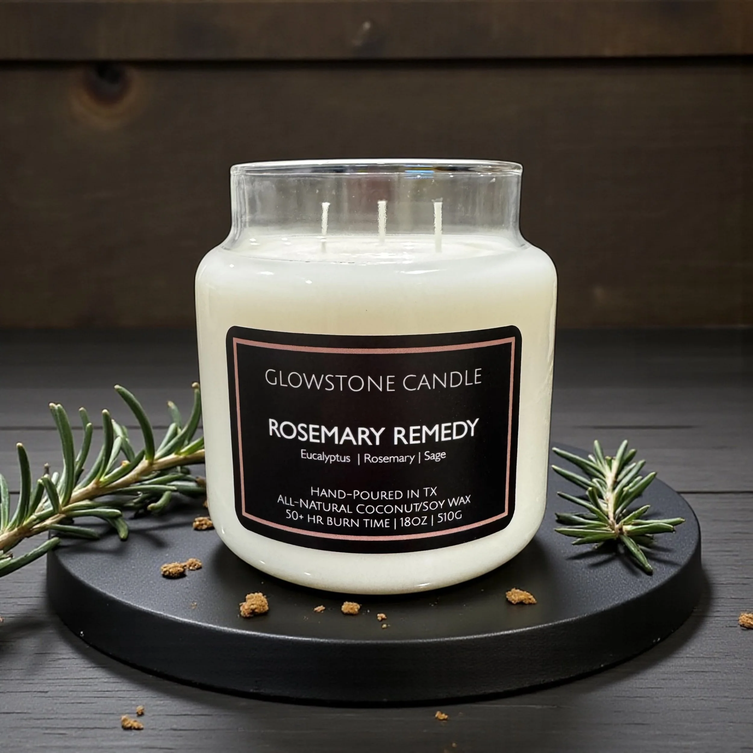 A photo of  the rosemary remedy candle in 18oz apothecary jar, white Wax and a black label.  The candle is on a wooden coaster with rosemary sprigs