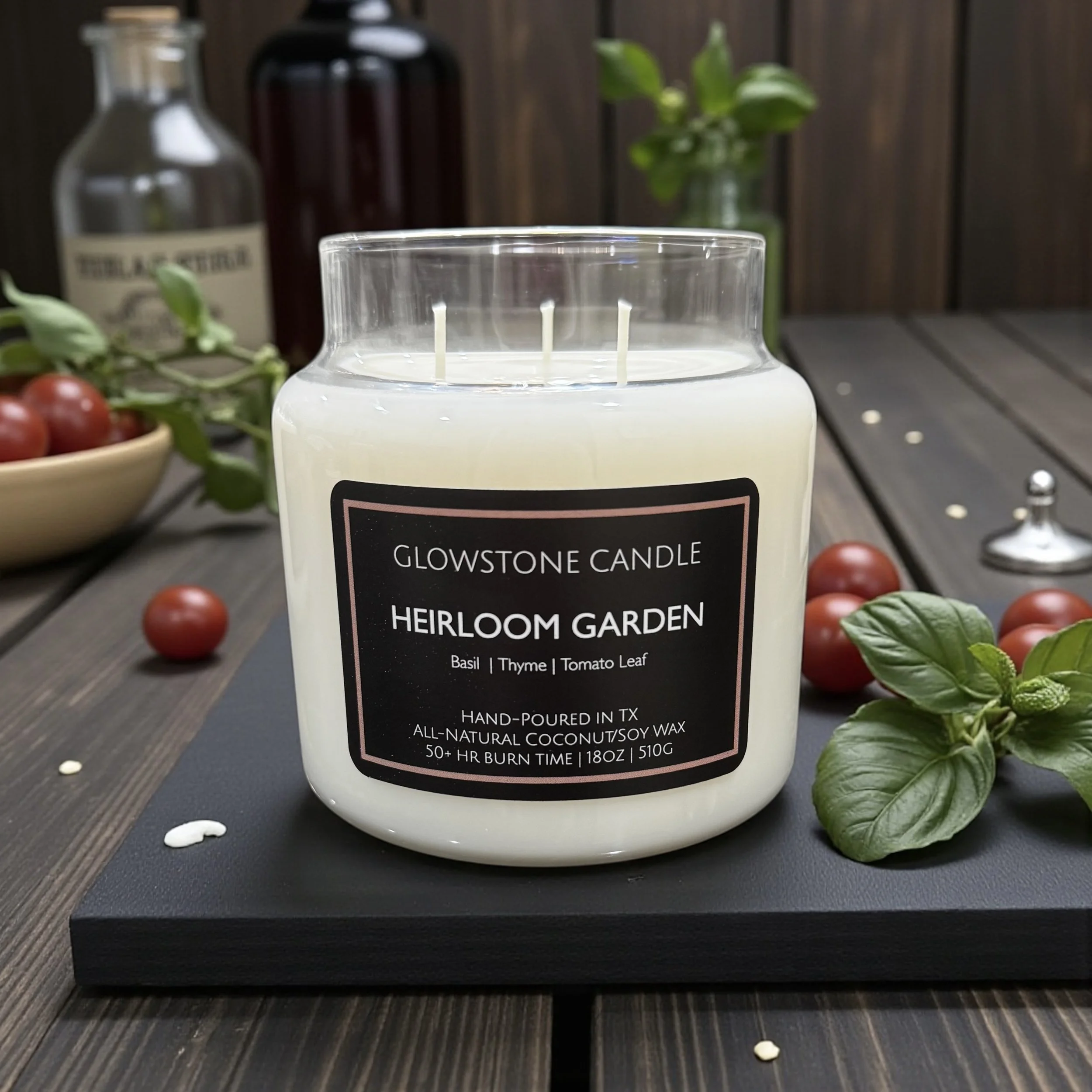 a photo of the heirloom garden candle showing its size of 18oz in an apothecary jar with white Wax and a black label. The photo also shows tomatoes, basil and apothecary bottles for aesthetic.