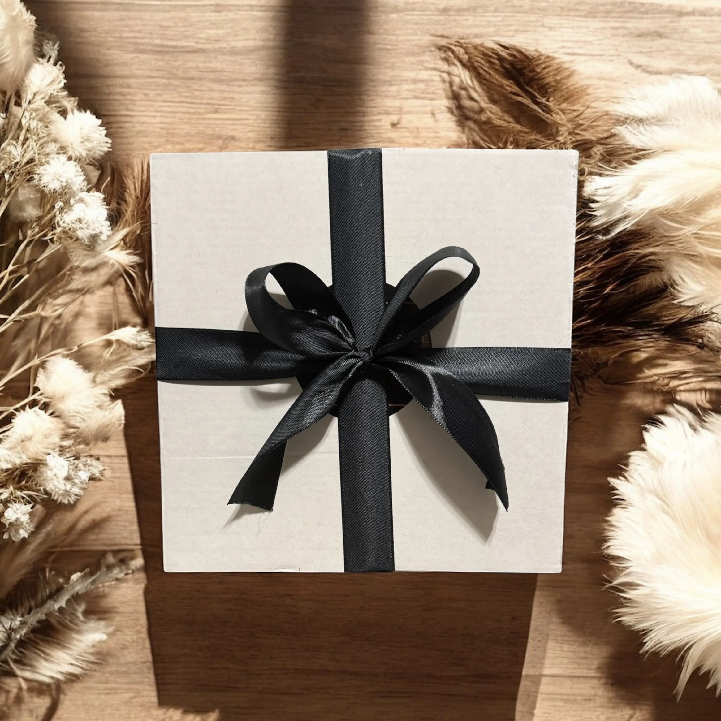 A white gift box with a black ribbon tied in a bow on top, placed on a wooden surface with dried white and beige foliage around it.