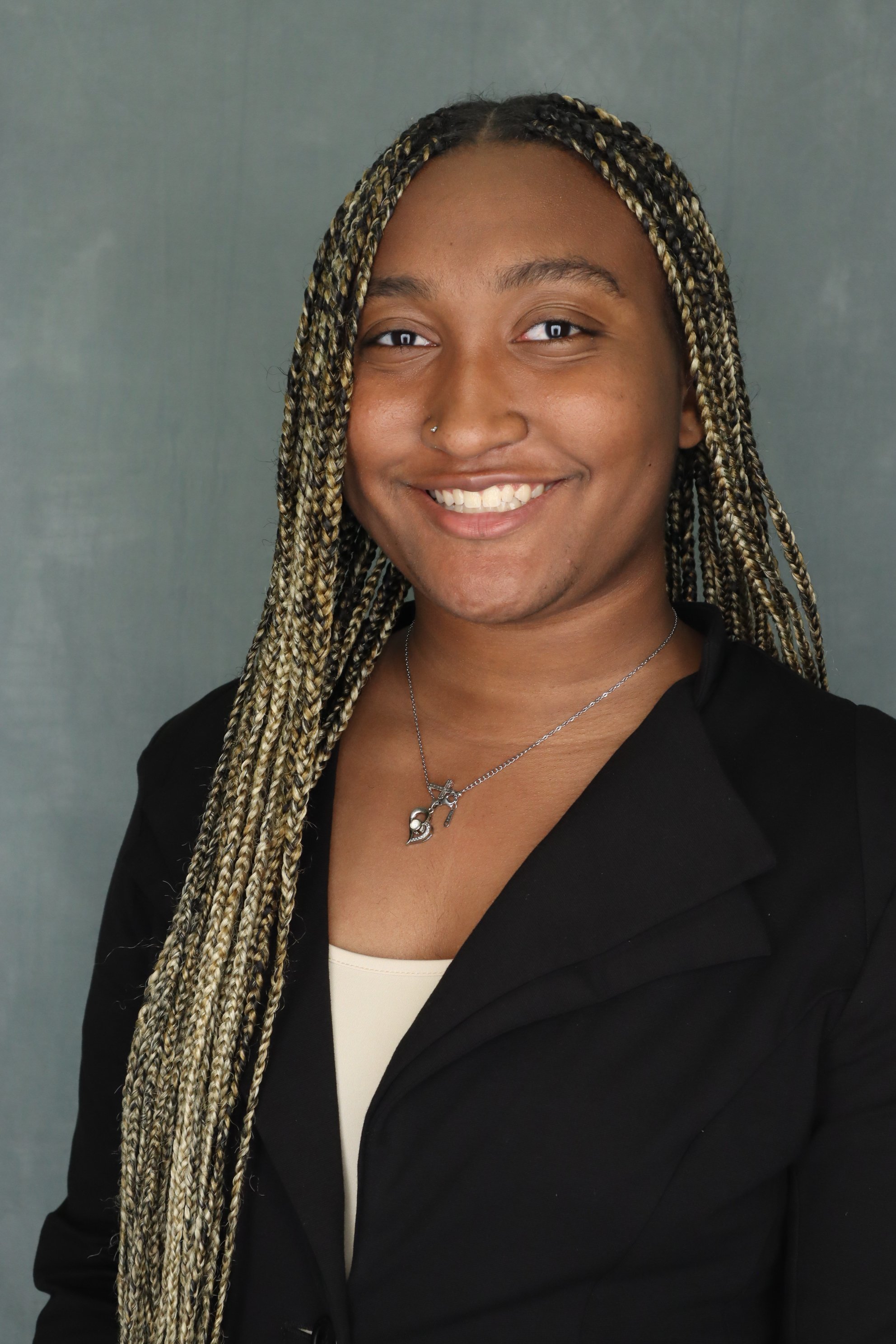 Portrait of a smiling woman with long blonde braids, wearing a black blazer, a cream top, and a necklace with a cat pendant, against a grey background.