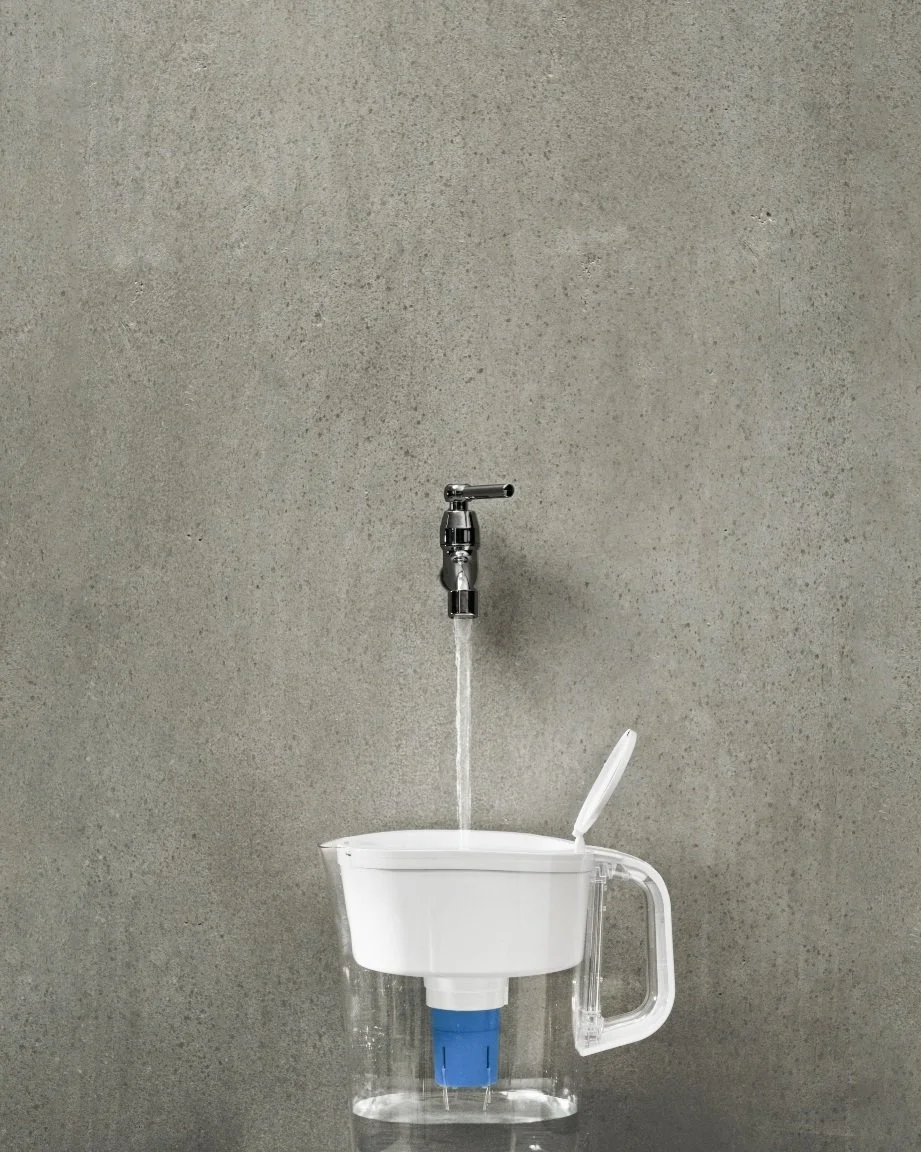 A water filter pitcher with a white lid and handle, placed under a faucet delivering water into it against a gray textured wall background.
