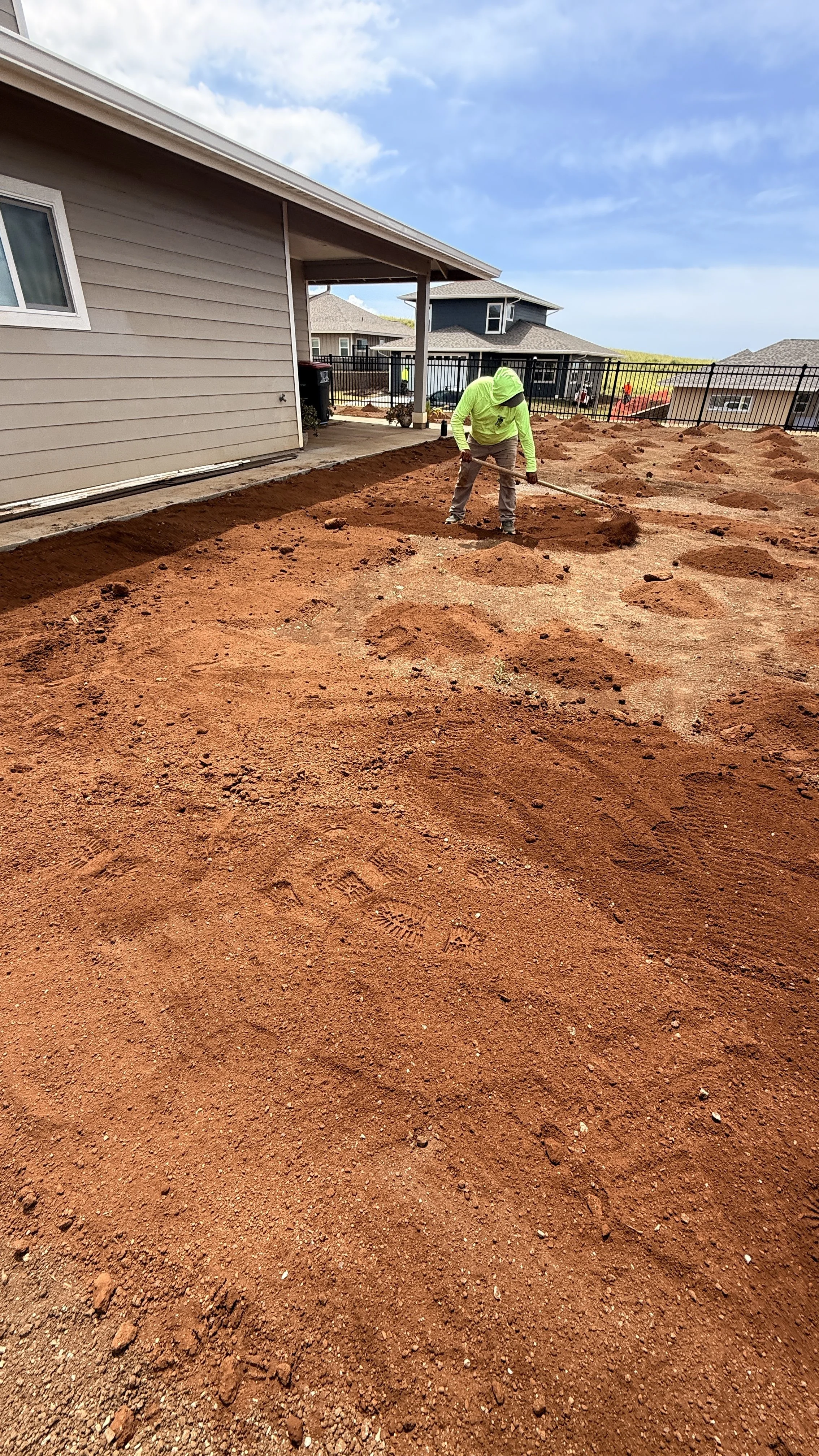 A worker leveling red soil with a rake in a backyard under a blue sky.