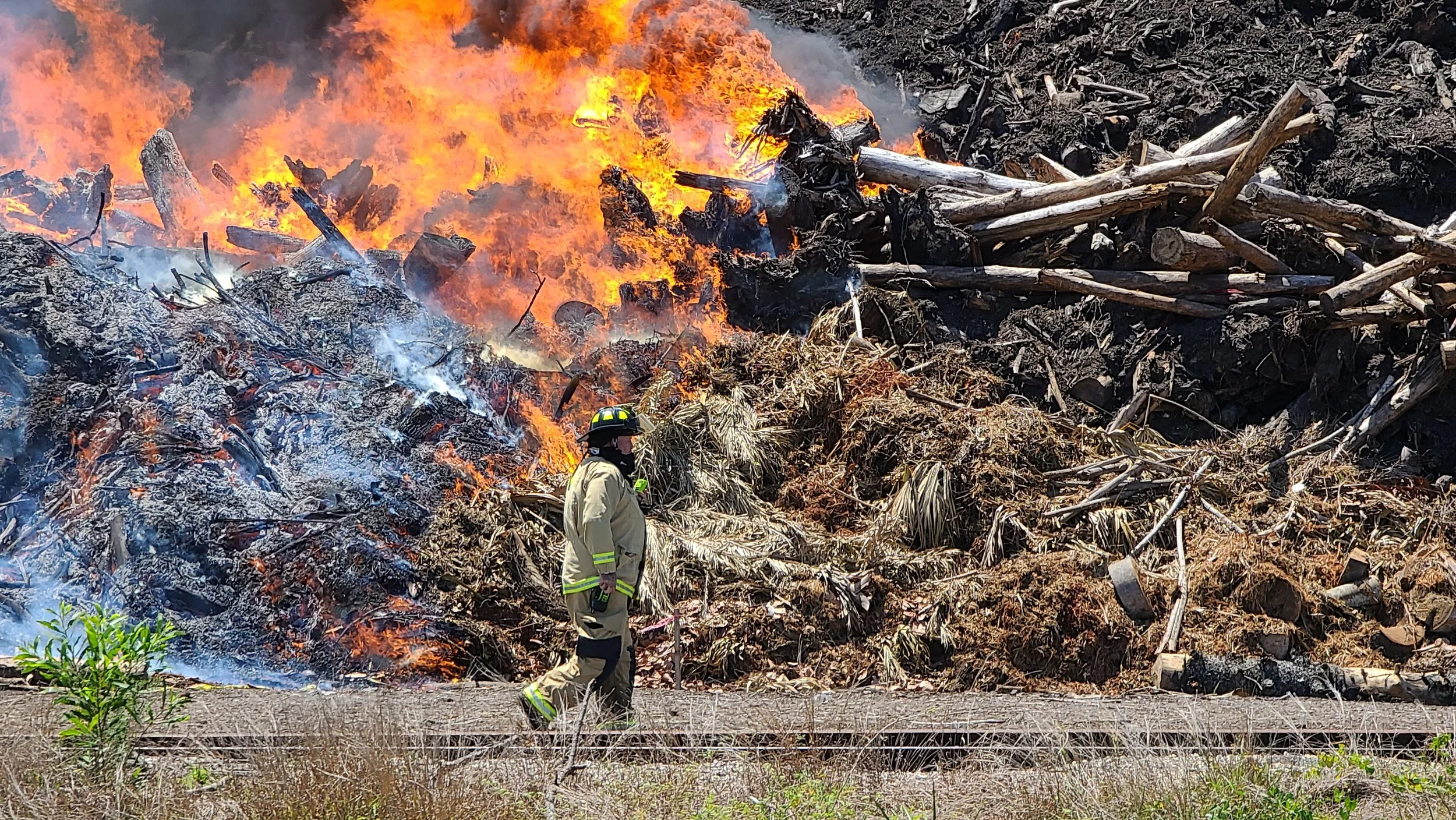 A wildfire burning through a dry, grassy area with flames and smoke. A firefighter in protective gear is standing in front of the fire, observing the scene.