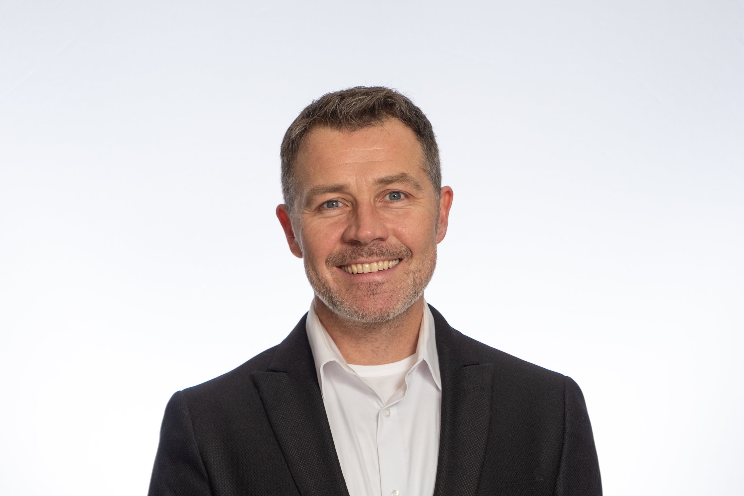 Portrait of Daniel Omar Frimannson, Axelyf's Head of Nucleic Acids and Biology; a smiling middle-aged man with short dark hair and a beard, wearing a black suit jacket and white shirt, standing against a plain white background.