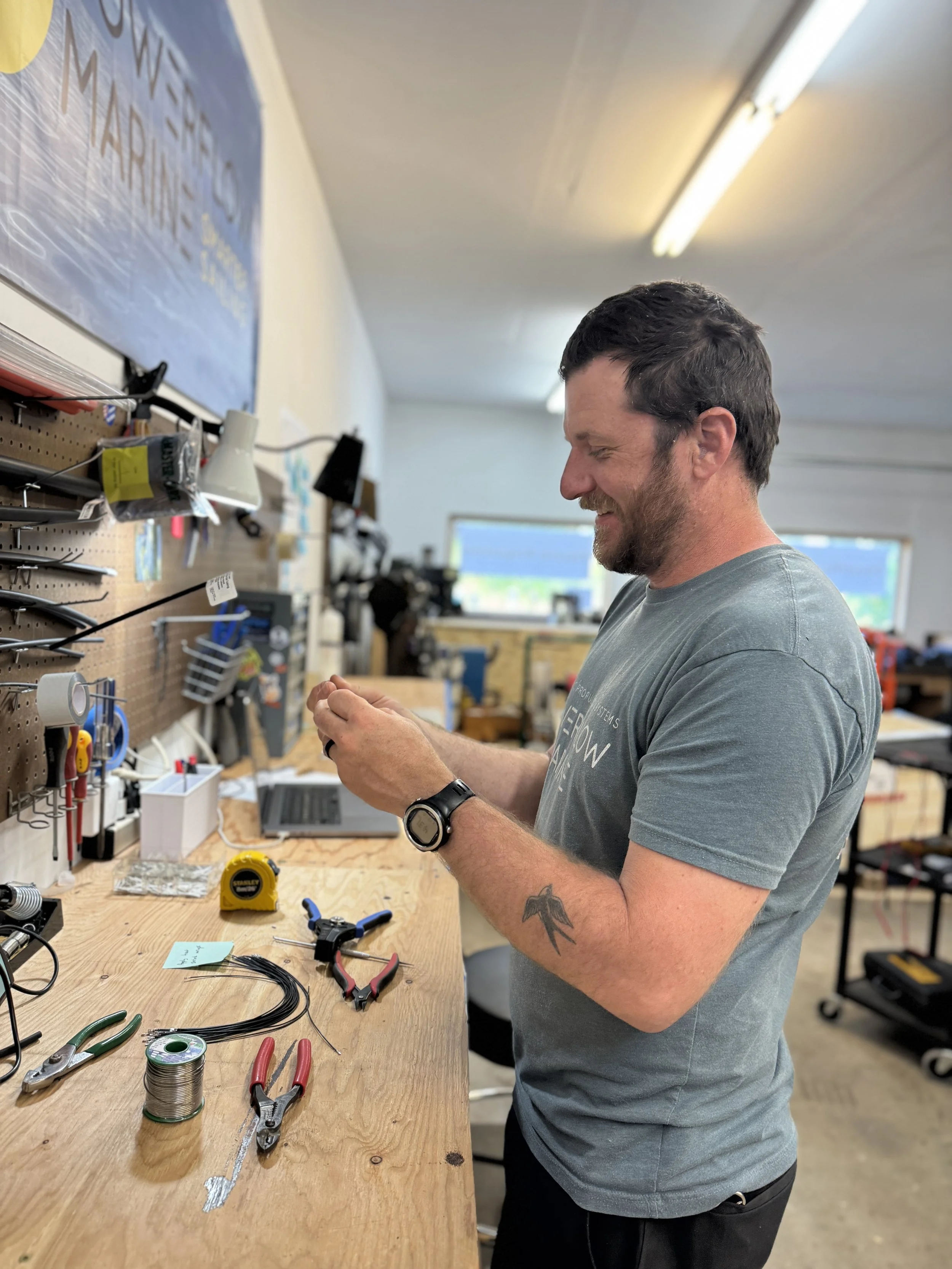 Smiling man working on electronics at a cluttered workbench in a workshop.