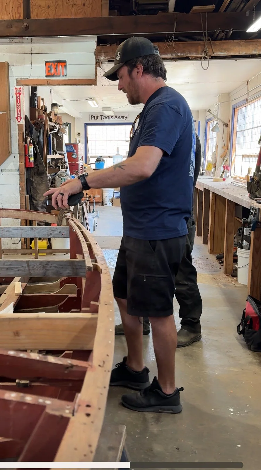 Man working on boat frame in workshop, wearing a cap, navy T-shirt, shorts, and sneakers.