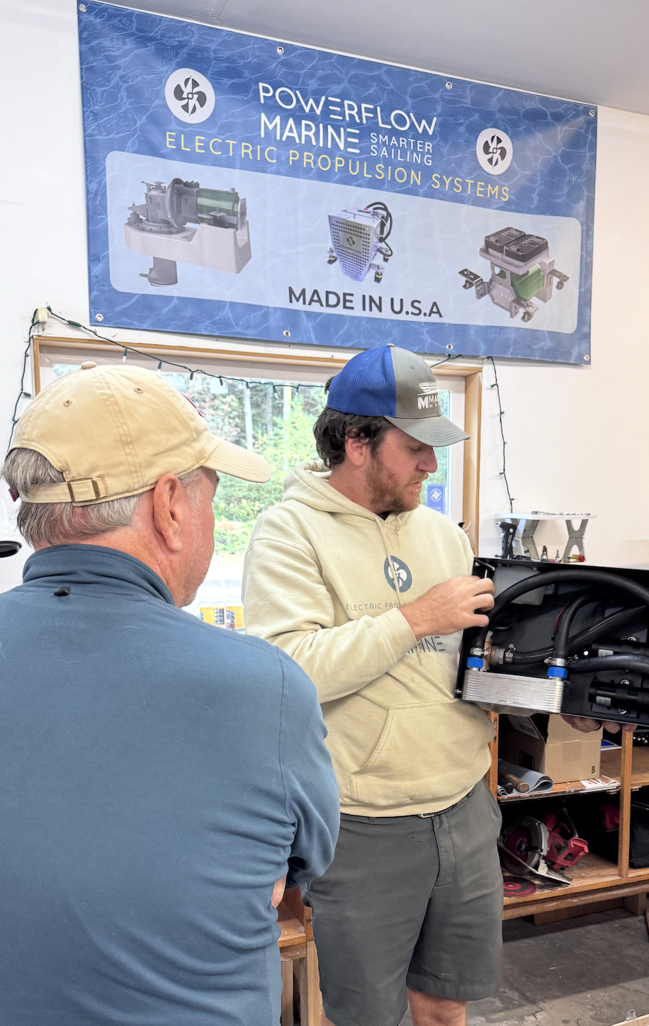 Two men in a workshop, one showing the other a black electric propulsion system component, with a banner above that reads 'Powerflow Marine, Electric Propulsion Systems, Made in U.S.A.' and images of marine electric propulsion products.