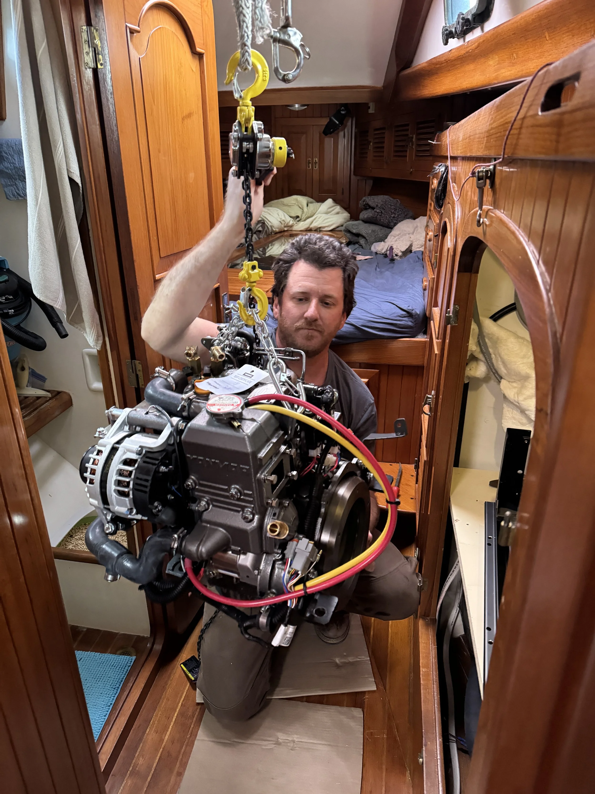 A man inside a small wooden room holding a boat engine and a hoist. The background shows a bed with unmade blankets and some pillows.