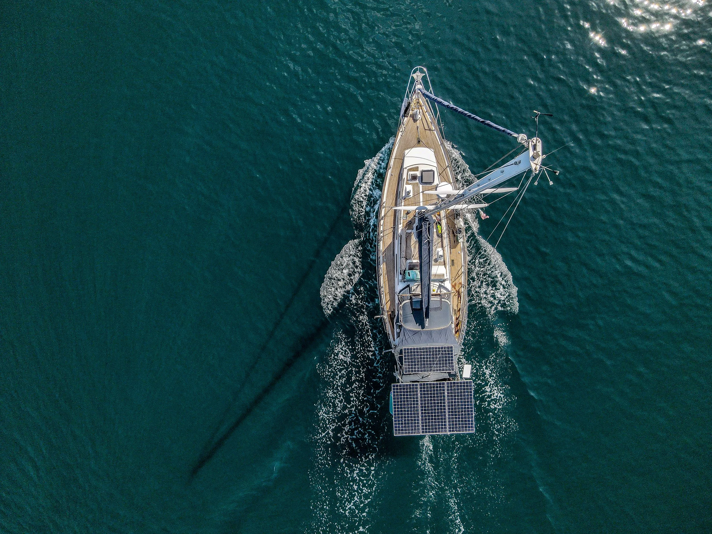 An aerial view of a sailboat with solar panels on the stern, moving through calm blue water.