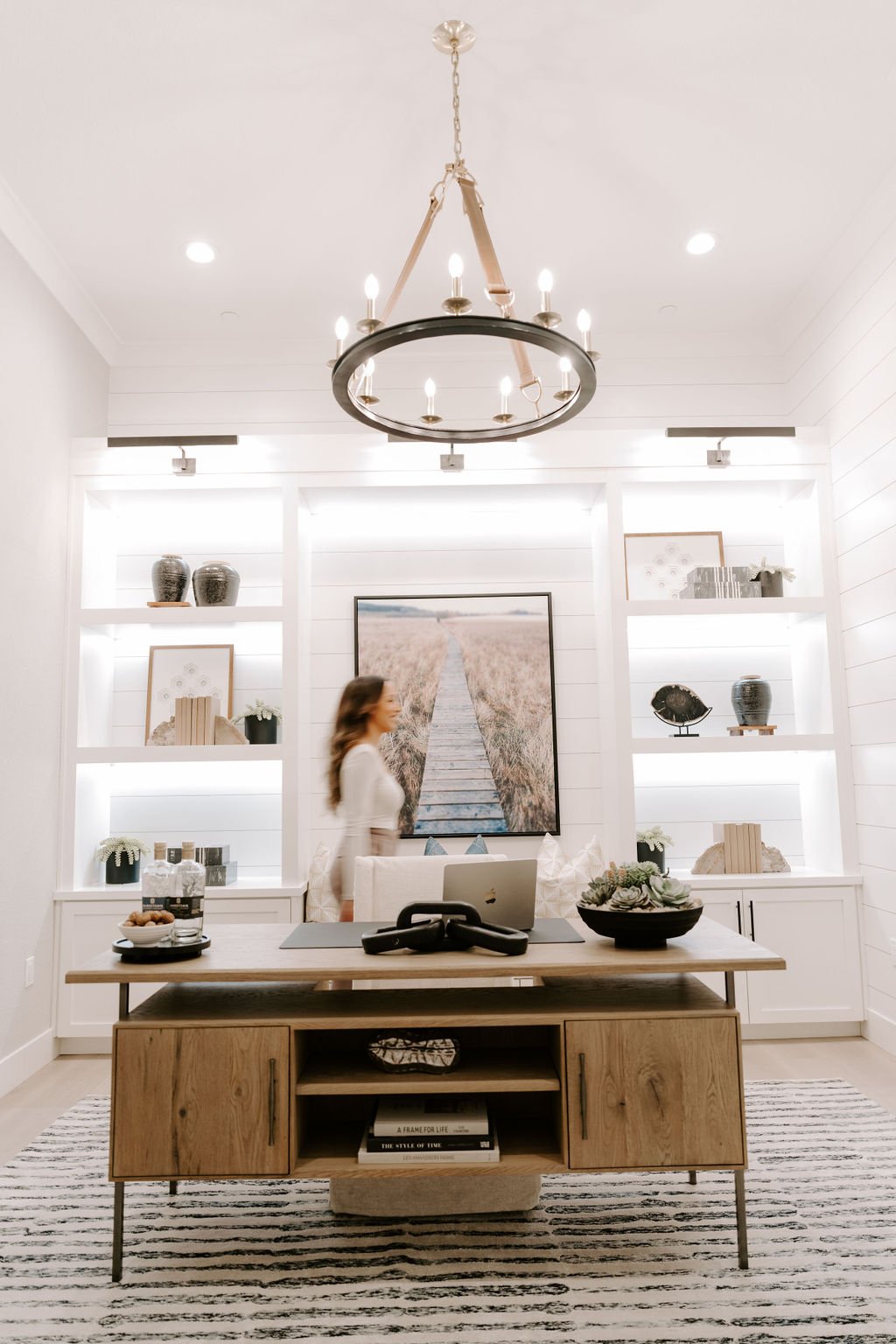 Modern living room with white walls, a wooden table with decorative items, a large wall art of a pathway through a field, built-in shelves with decorative objects, and a chandelier hanging from the ceiling.