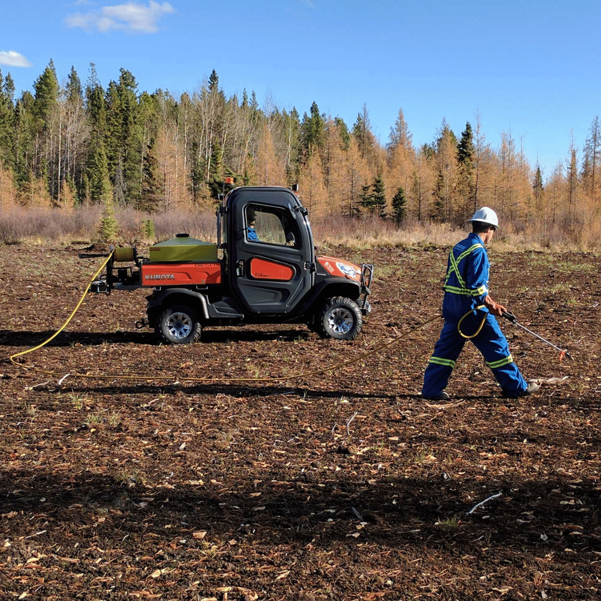 Man standing beside a UTV with hose reel, spraying in the brush for land reclamation under blue skies in Alberta.