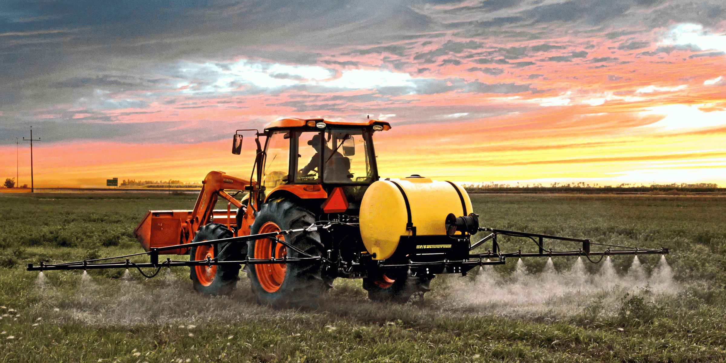 Wide-angle shot of an agricultural boom sprayer operating on a orange tractor in a lush green field in Alberta.
