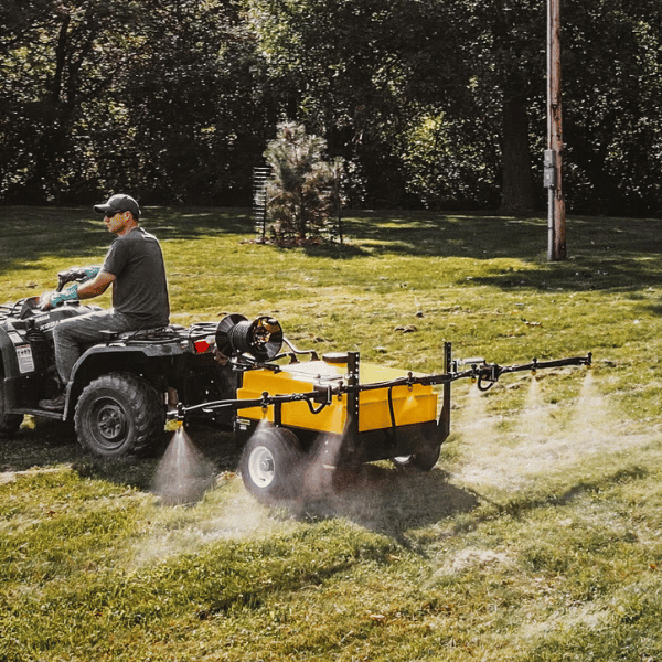 Side view of a 50-gallon Pull-Behind Mini Sprayer in an agricultural yard, with a homeowner on a quad in Alberta.