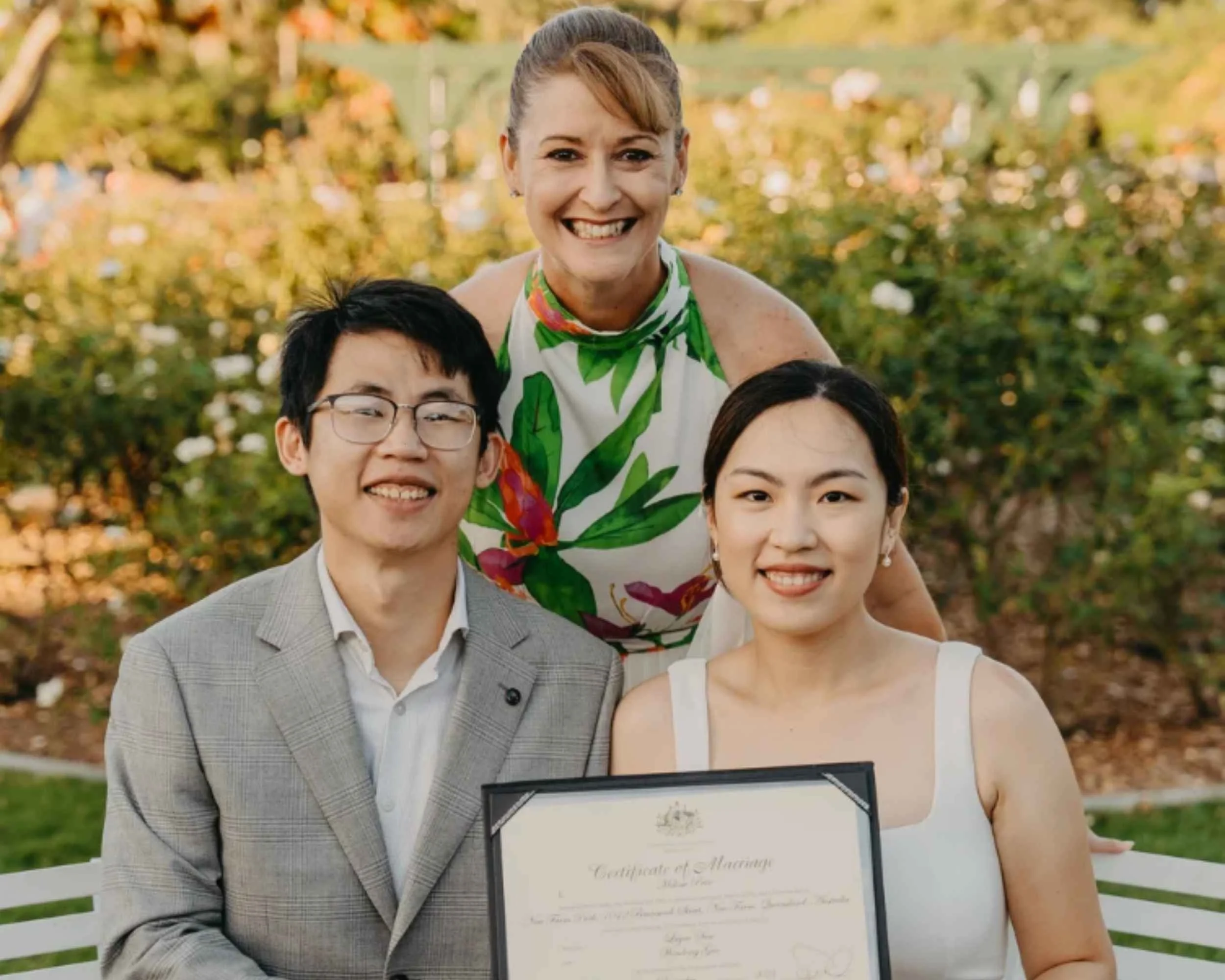 A couple holding a marriage certificate with a woman standing behind them, smiling. The couple is seated at a white outdoor table with bouquets of flowers, surrounded by a garden setting.