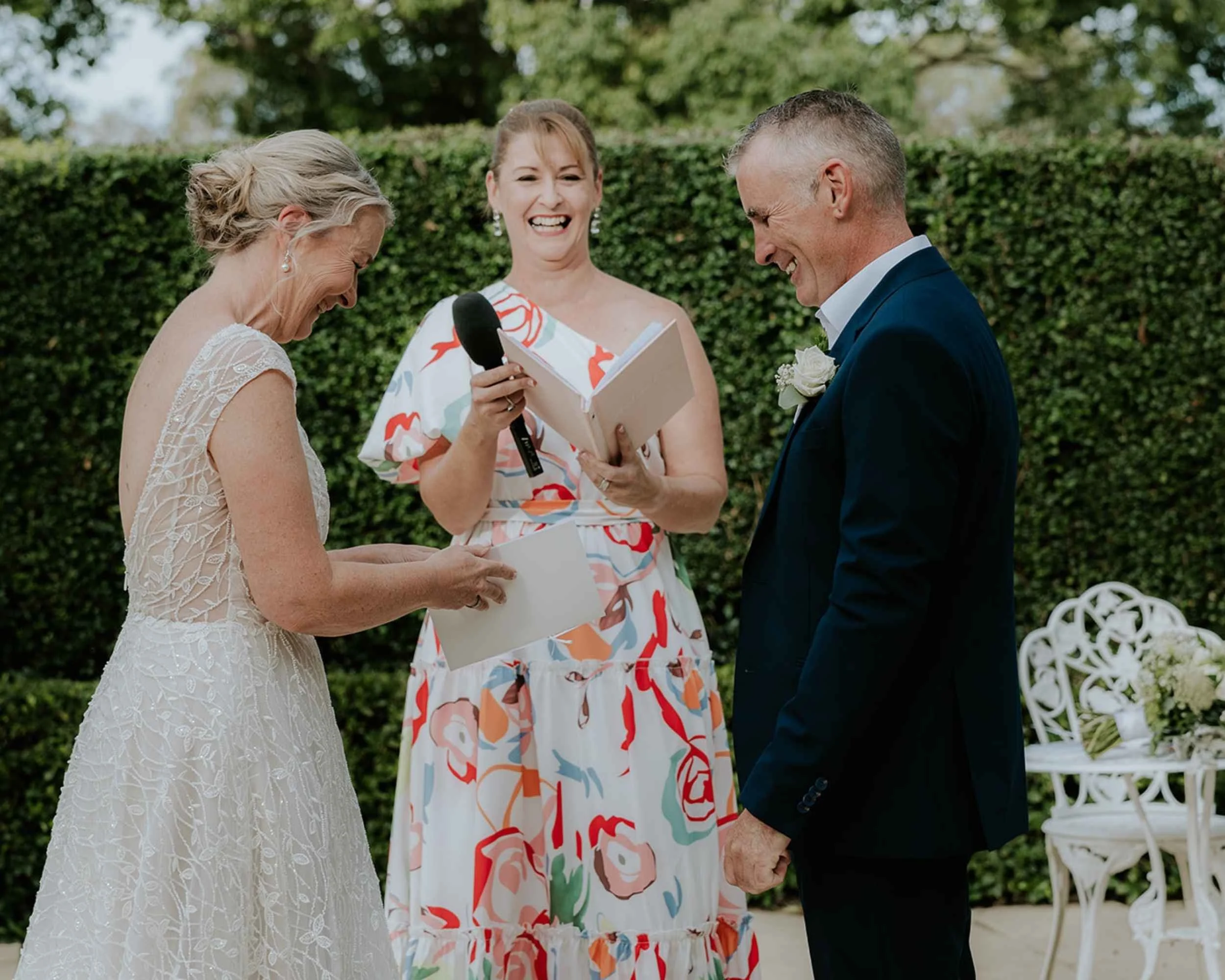 Bride and groom smiling during wedding ceremony with officiant holding microphone and book, outdoor garden setting.