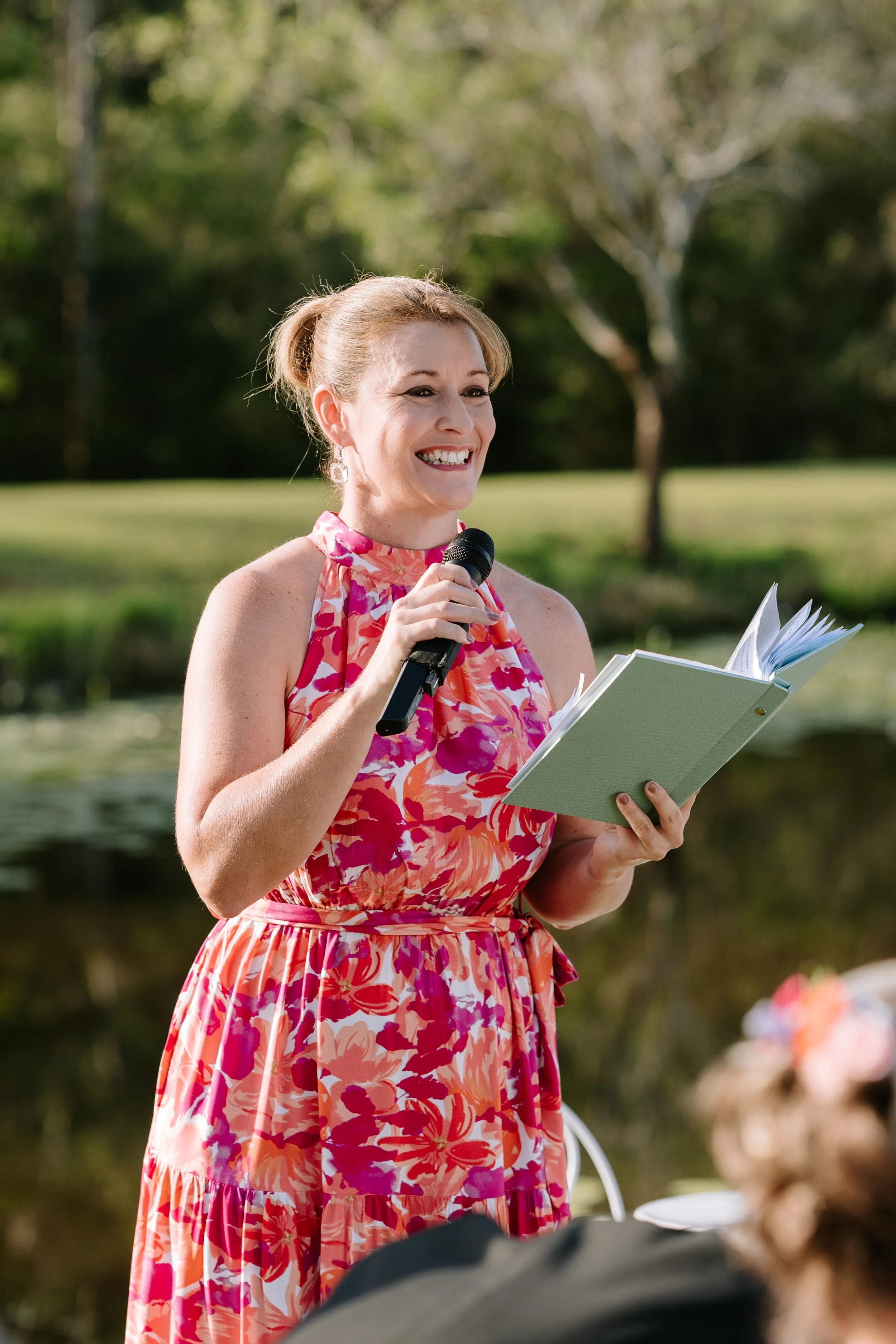 A woman in a colorful floral dress holding a microphone and a book, smiling outdoors.