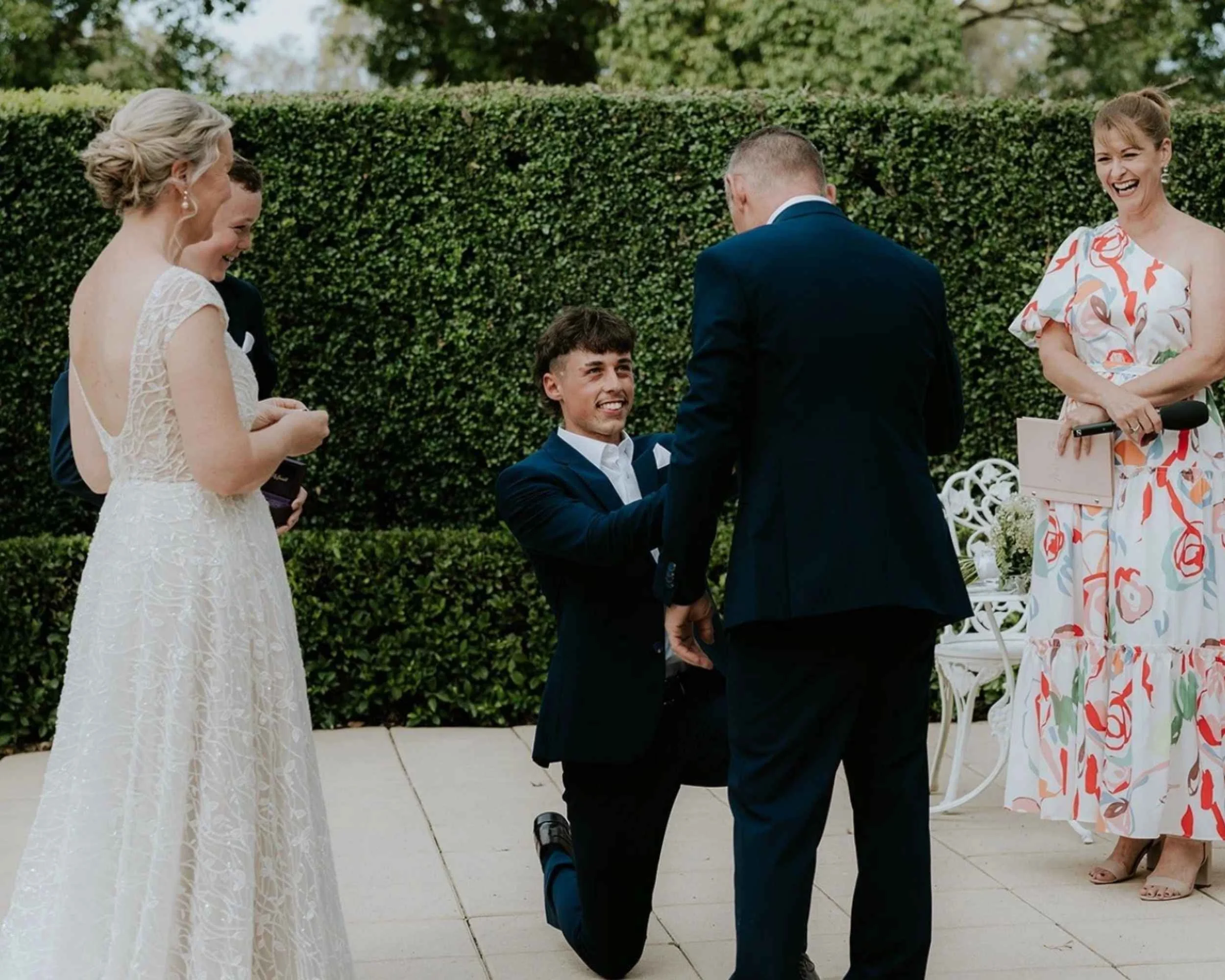 A group of people at an outdoor wedding ceremony, featuring a person in a wedding dress, two individuals in formal suits, one of whom is kneeling, and a person in a floral dress holding a microphone.