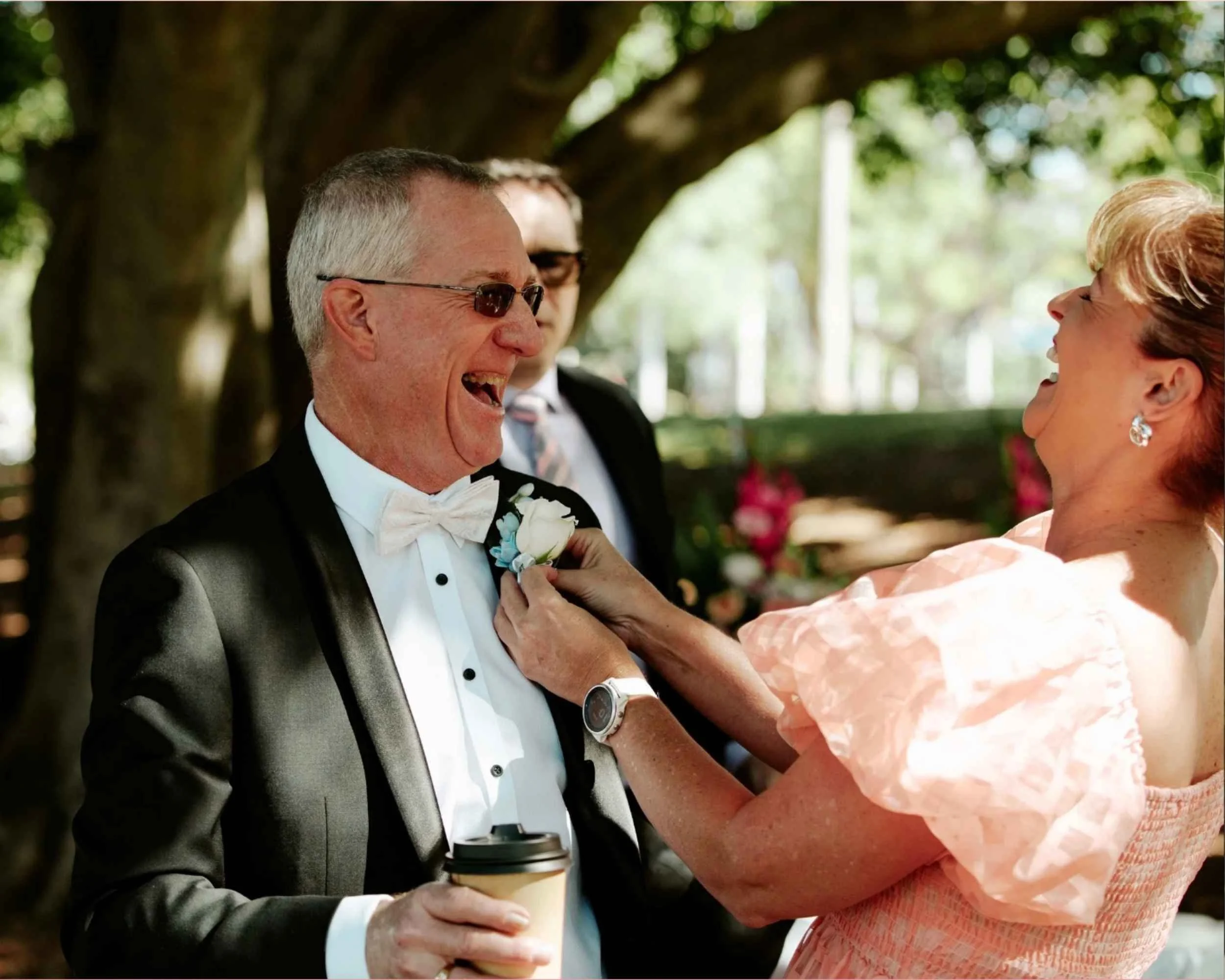 A man in a tuxedo, laughing, holding a coffee cup, while a woman pins a boutonniere on his jacket. They are outdoors with trees in the background.