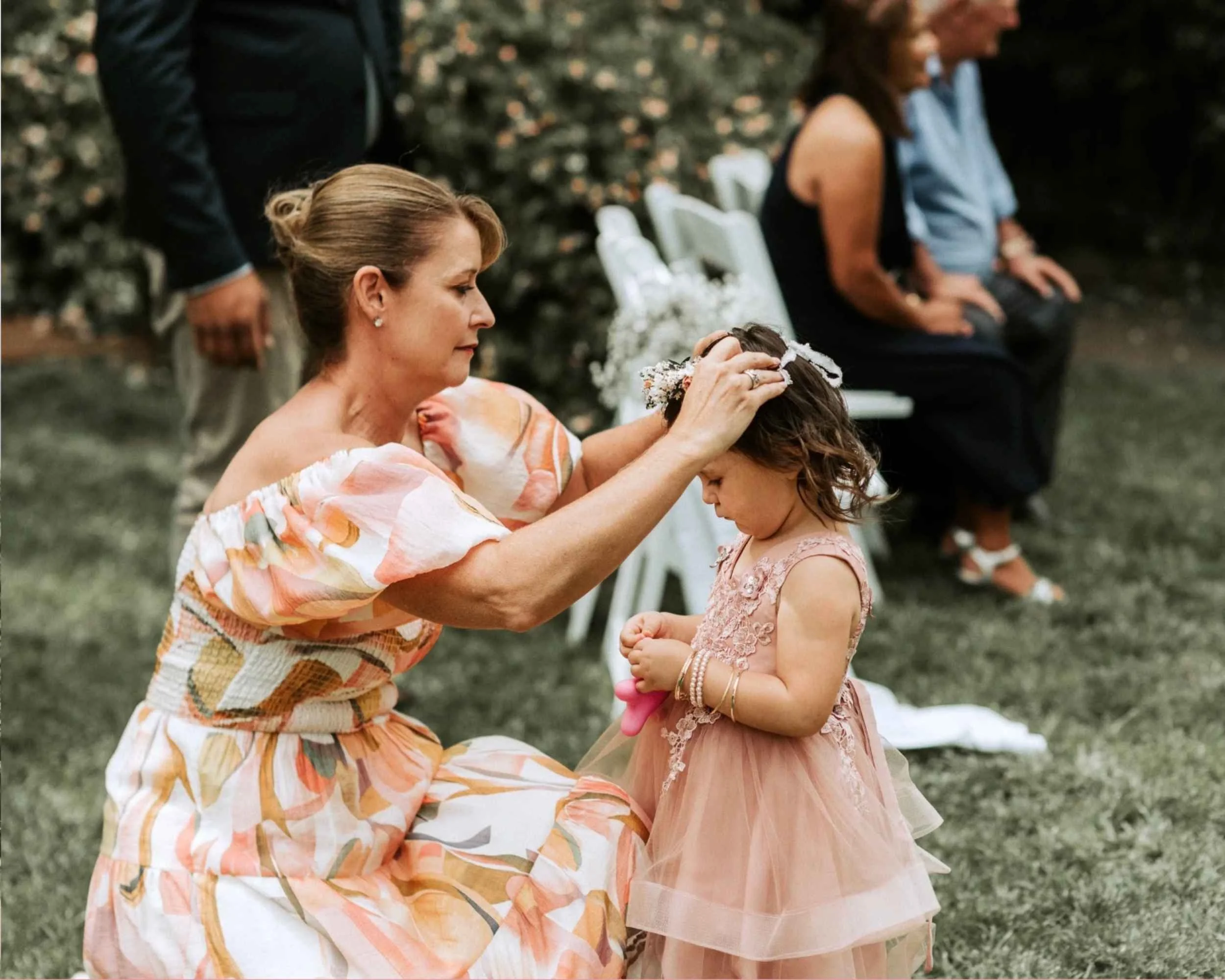 A woman in a floral dress adjusting a flower crown on a young girl in a pink dress. They are outdoors, with seated guests blurred in the background.