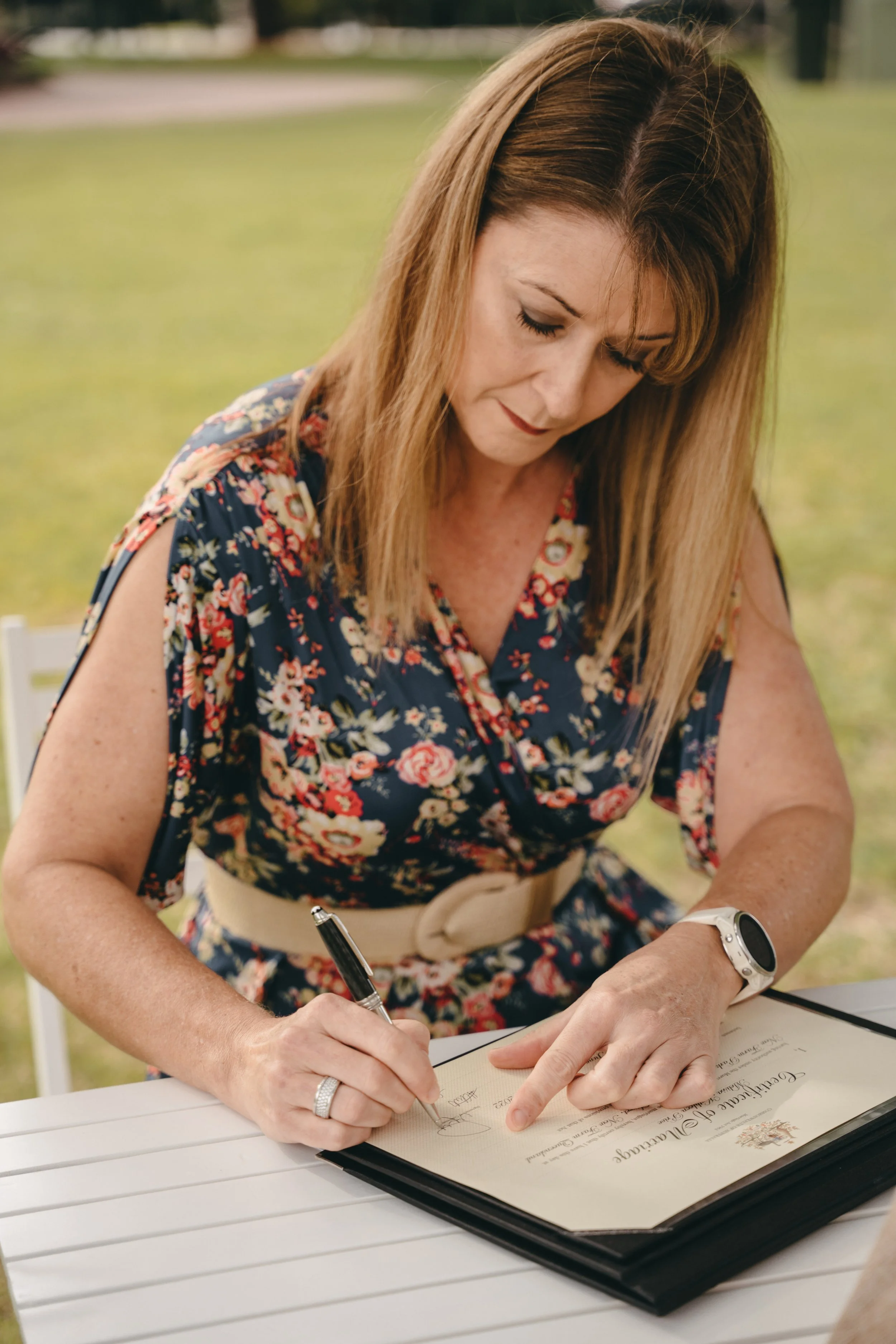 Woman signing a certificate outdoors wearing a floral dress and a smartwatch.